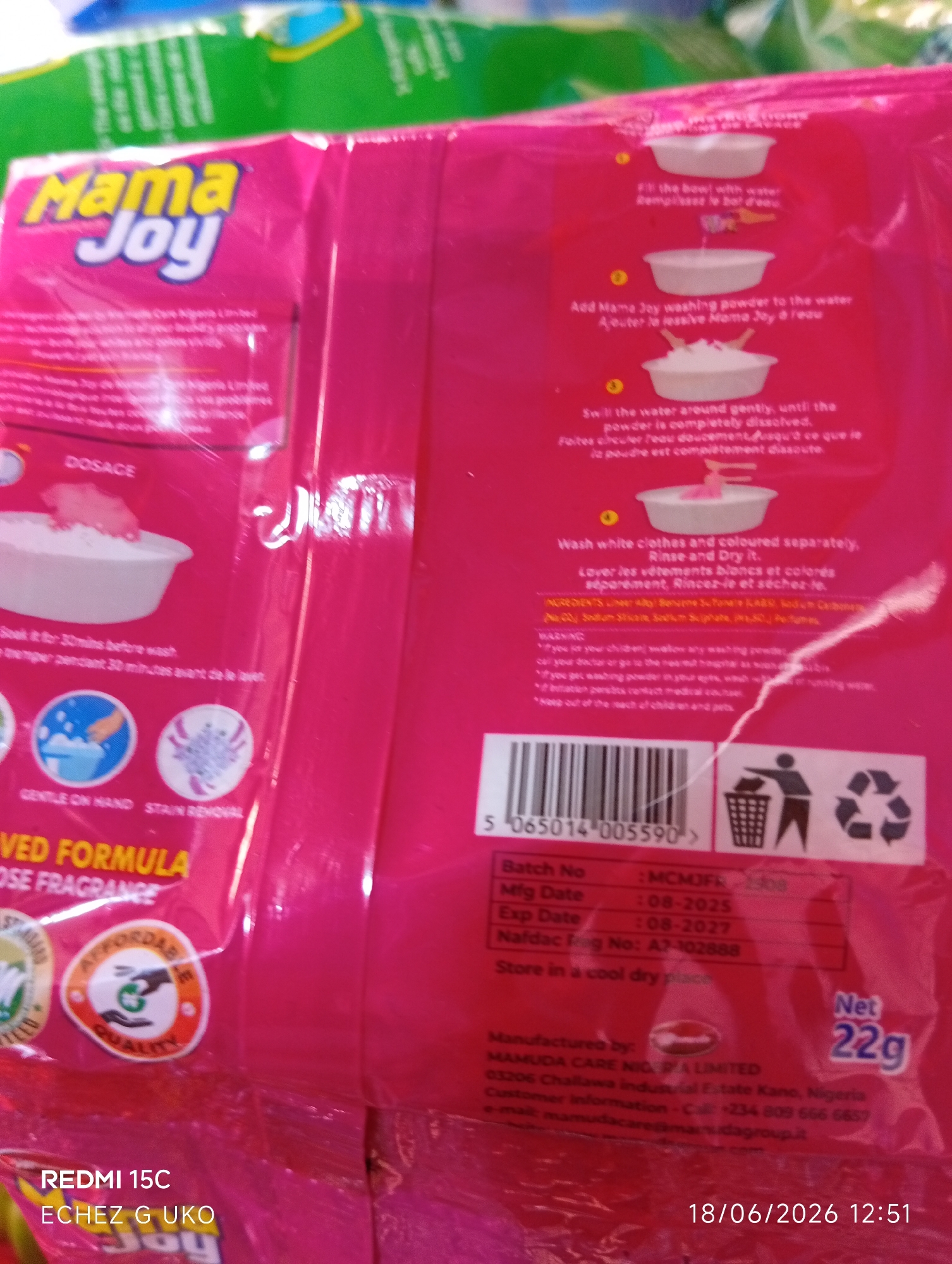 The image shows a close-up of a pink package of Mama Joy washing powder. The package features the brand logo prominently at the top. There are instructions on how to use the product, illustrated with simple drawings. Text on the package is in both English and French, indicating usage instructions like "Fill the bowl with water," "Add Mama Joy washing powder to the water," and "Swill the water around gently, until the powder is completely dissolved." It also mentions "Wash white clothes and coloured separately, Rinse and Dry it."
Below the usage instructions, there are icons indicating "Gentle on Hand" and "Stain Removal." Further down, text reads "VED FORMULA" and "OSE FRAGRANCE." Two circular emblems are visible: one with "AFFORDABLE" and "QUALITY" and another with a leaf design.
At the bottom of the package, there is a barcode, followed by product details like "Batch No," "Mfg Date," "Exp Date," and "Nafdac Reg No." The manufacturer is listed as "MAMUDA CARE NIG. RA LIMITED" with an address in Kano, Nigeria. Contact information, including a phone number and email address, is also provided.
The bottom right corner of the image displays a date and time stamp: "18/06/2026 12:51". The background is blurry but suggests other products or packaging, possibly in a retail environment. The overall impression is of a product being presented for sale or information.