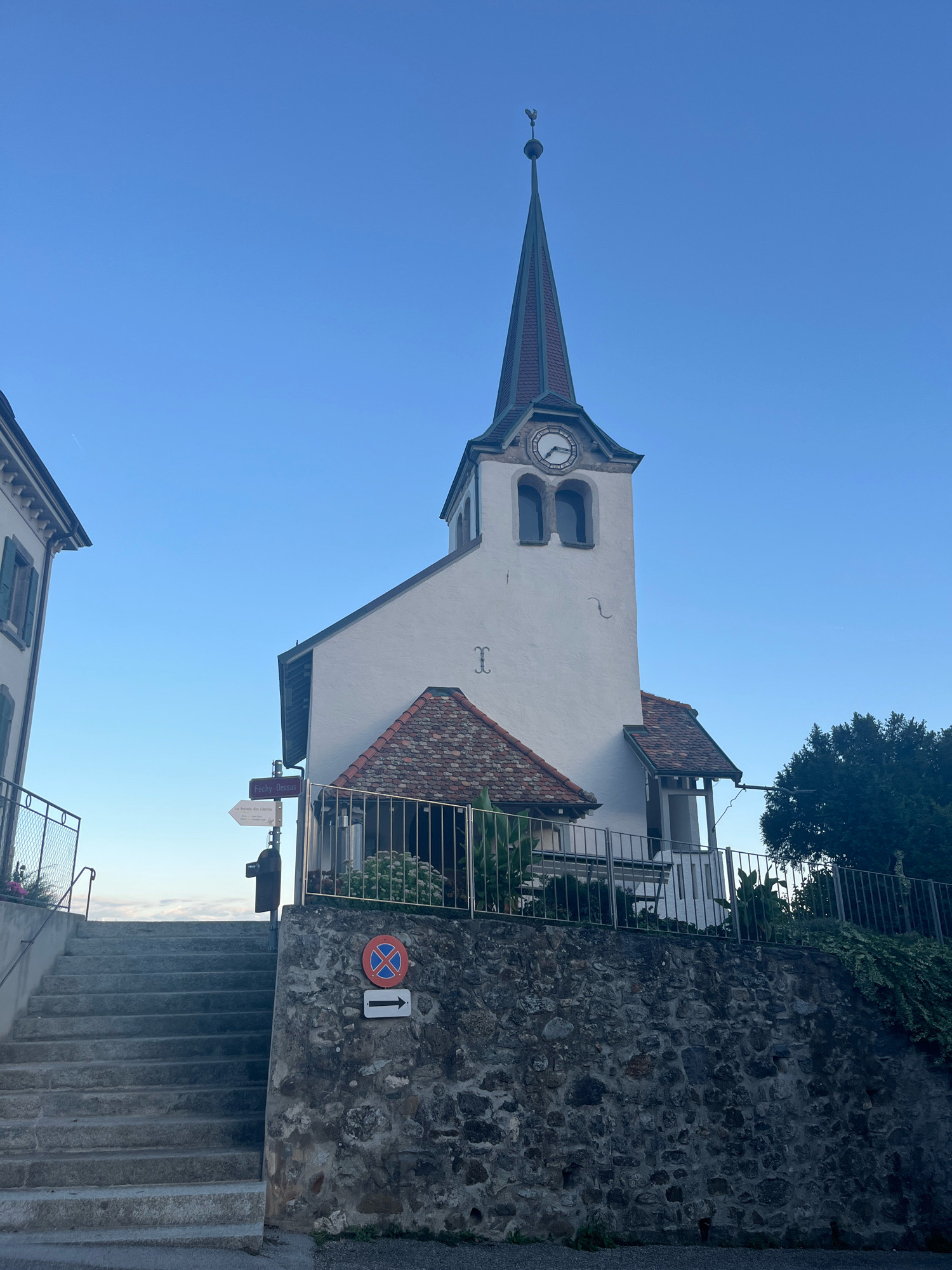 The image shows a white church with a tall, narrow spire topped with a weather vane. The church has a clock face on its tower and arched windows. The building is situated on a stone wall, with a set of stone stairs leading up to it from the left. A metal fence encloses the area around the church. There are directional signs and a traffic sign posted on the wall near the stairs. In the background, there is a clear blue sky and some trees. The scene appears to be in the town of Féchy, Switzerland, based on the directional sign which reads "Fechy-Dessus" and "La Suisse des Capites". There are no people visible in the image. The lighting suggests it is either early morning or late afternoon.