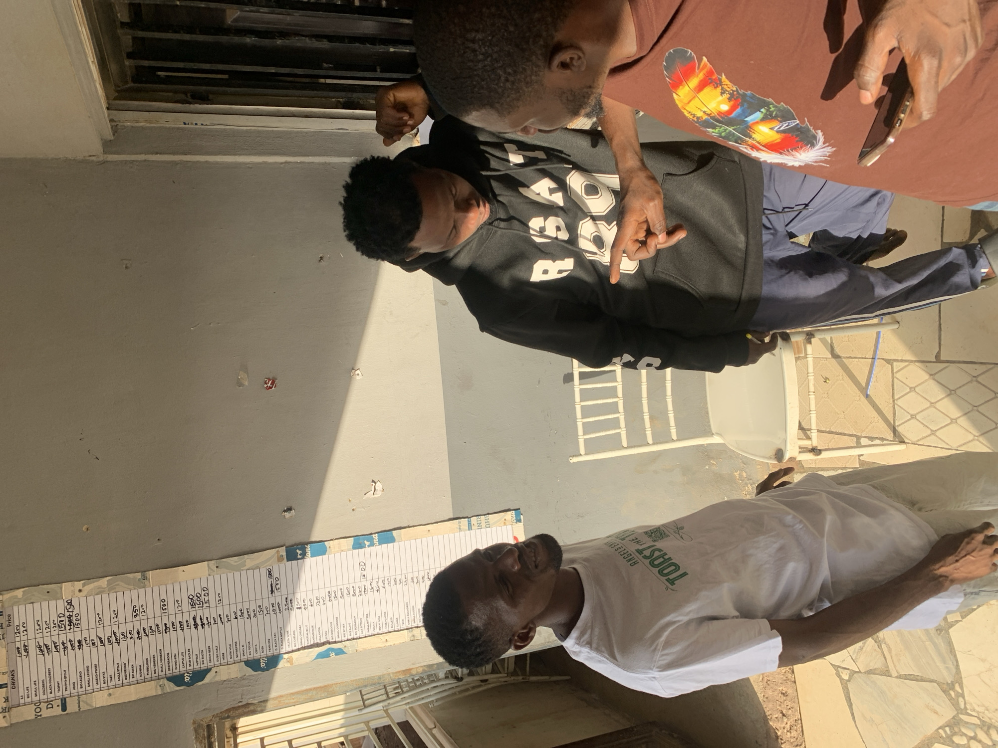 Three adult males are observed interacting in an outdoor, tiled area in Kabong, Nigeria, adjacent to a light gray wall.
On the left, a man with dark skin, a short beard, and short dark hair wears a white t-shirt with green text and graphics, and light-colored trousers. He is facing right, smiling, and observing the other two men. His left arm is visible, showing prominent veins.
In the center, a man with dark skin and short dark hair wears a black hoodie with "RBSAT" and "89" printed in white on the front, and dark blue track pants with white side stripes. He is looking downwards and to his right, with his hands near his waist. He wears a dark slide sandal on his right foot.
On the right, a man with dark skin and short dark hair/beard wears a brown t-shirt featuring a colorful feather graphic depicting a sunset landscape. He holds a light-colored smartphone in his left hand. His right index finger is pointed towards the man in the black hoodie, indicating an explanatory or directive gesture. He is looking towards the man in the black hoodie.
A long, vertical paper price list, labeled "DRINKS" and "Price," is taped to the light gray wall on the left. It displays various drink names and prices, some of which are crossed out and updated (e.g., "BIG LIFE 1200," "TROPHY 1500," "BUDWEISER 1300," "STAR RADLER 1500"). To the right of the price list, the wall shows several small, faded red and white paper fragments.
A white plastic chair with a simple metal frame is positioned behind the man in the white t-shirt. The floor consists of irregular light-colored stone tiles on the left and darker, square-patterned tiles on the right. To the far right, a section of vertical metal security bars is visible. Strong natural light illuminates the scene, casting distinct shadows.