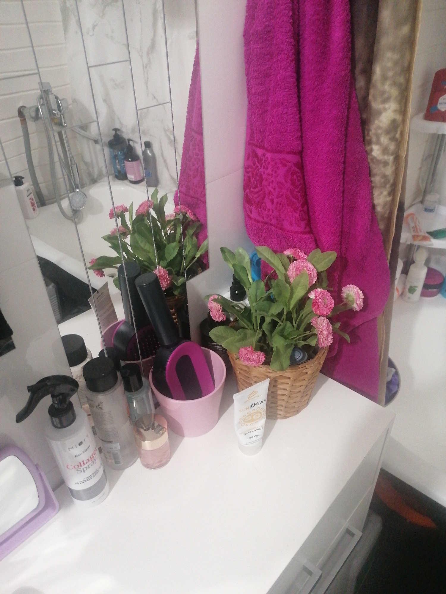 A high-angle, medium shot captures a cluttered bathroom countertop with various personal care items. In the foreground, a white spray bottle labeled "Collagen Spray" with "Hair Expert" and other text is visible. Next to it are several other bottles, a small pink jar, and a tube of "SUN CREAM" with "SPF 50" printed on it. To the right, a small potted plant with pink flowers sits in a woven basket. Behind the plant and bottles, a large, bright pink towel is draped over something out of frame. A large mirror reflects the scene, showing a shower head and faucet, more bottles, and some of the pink towel. The countertop appears to be white, and the wall behind it is tiled with a marble pattern. The floor is dark and reflects light. The overall impression is of a well-used, functional bathroom space, possibly in a residential setting in Angarsk, Russia, given the context provided. There are no people visible in the image.