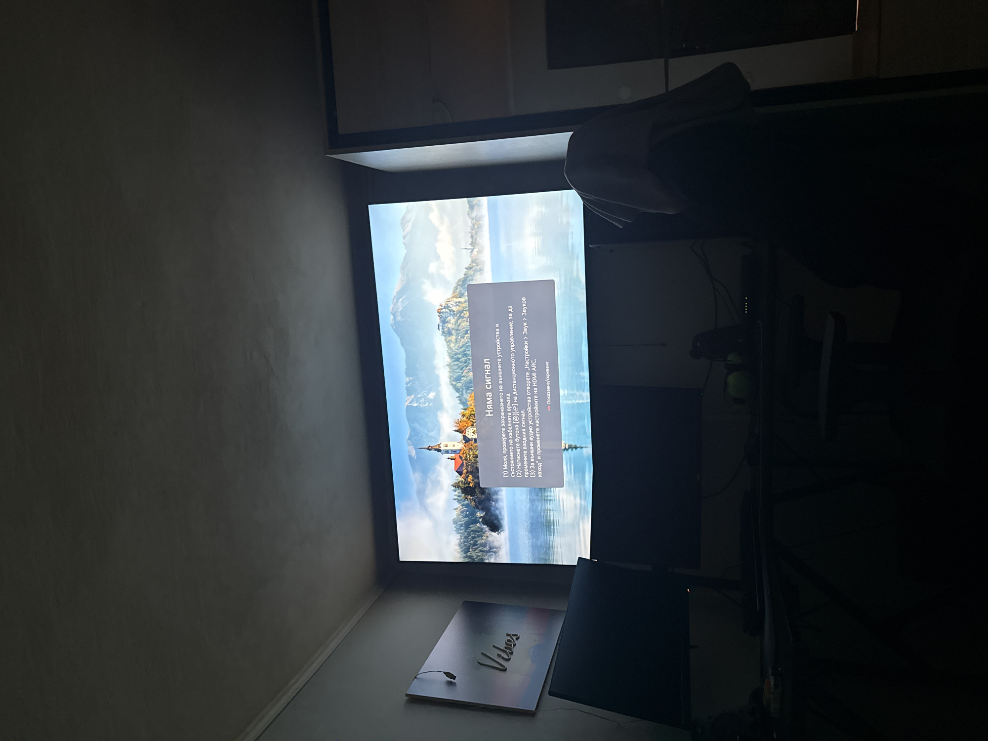 Here's a description of the image:
The image shows a dark room with a TV screen displaying a scenic view of a lakeside town with mountains in the background. A message on the screen indicates "No Signal" in Bulgarian, with instructions to check connections and settings. Below the TV, there's a lighted sign that says "Vibes." The rest of the room is in shadow, with a desk and other objects faintly visible to the right of the TV. There is nothing in the photo that indicates a location in the city of Shumen, Bulgaria.