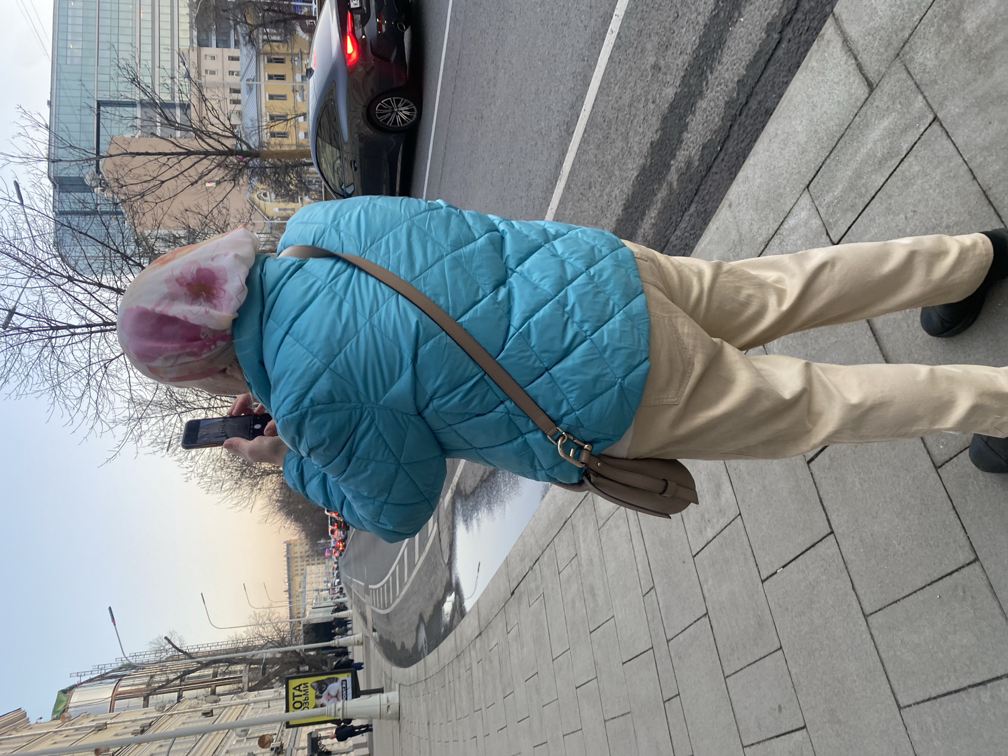 A person is depicted from behind, standing on a grey paved sidewalk and holding a black smartphone horizontally with both hands, aimed forward as if capturing an image. The individual wears a diamond-quilted teal puffer jacket, light beige trousers, and black shoes. A tan leather cross-body handbag with metallic hardware is draped over their right shoulder. A white headscarf, patterned with pink and red floral motifs, covers the person's head.
The setting is an urban streetscape, identified as **Novy Arbat Street in Moscow, Russia**. The ambient light suggests late afternoon or early evening, with a pale blue sky transitioning to a faint warm hue at the horizon. The wide asphalt road comprises multiple lanes of traffic, separated from the sidewalk by a concrete curb. A puddle is visible in the road's shoulder. Bare deciduous trees line the street.
On the left side of the street, a sequence of multi-story buildings with light-colored facades is visible. One building exhibits a partially obscured Cyrillic sign featuring "ОТА" and "ТОЧКА" (likely referencing "Открытие" and "Точка" banks). Distant pedestrians are present on the sidewalk. Across the road to the right, a dark-colored coupe automobile is visible, its red taillights illuminated. The background features a blend of older architectural styles and a prominent, multi-story modern building with a glass facade. Red vehicle lights indicate distant traffic.