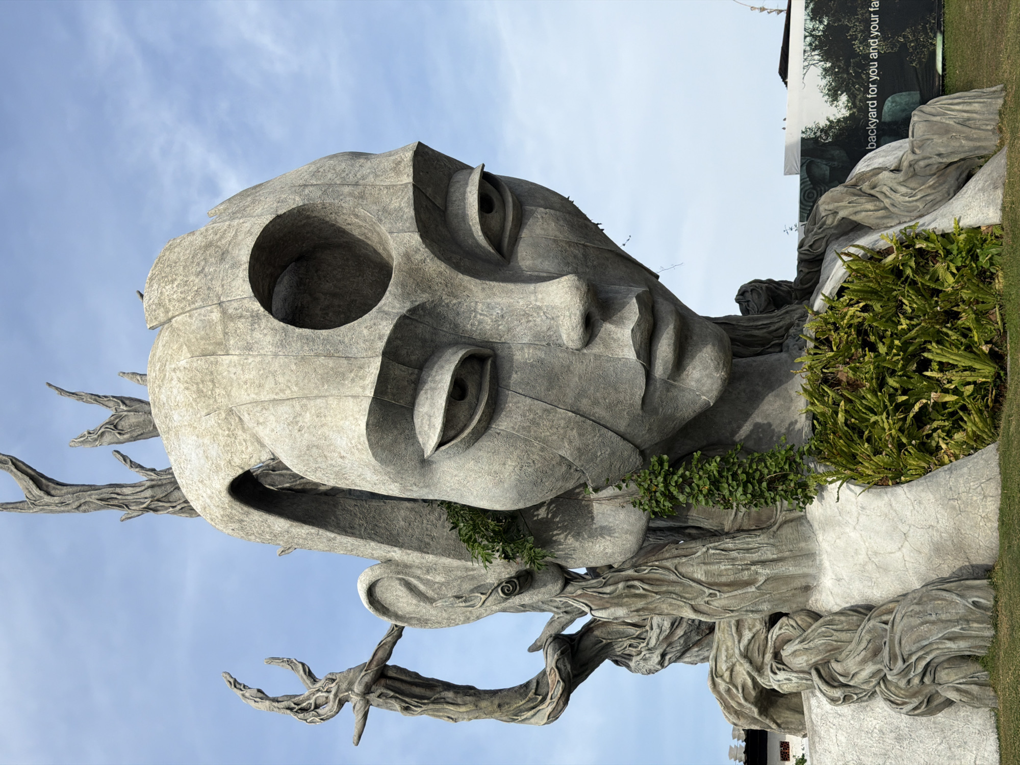 A large, stone sculpture of a stylized human head emerges from a landscape with a bright blue, partly cloudy sky. The sculpture's face is serene, with large, almond-shaped eyes and a slightly parted mouth. The head is partially covered by a rough, bark-like texture that suggests a fusion with nature, with some green vines growing around the face and ears. Atop the head, several abstract, branch-like forms protrude towards the sky. The sculpture is situated in an outdoor setting, with a patch of green grass at its base and a large banner visible in the background. The banner, with a dark, wooded image and white text, reads "backyard for you and your fam". This sculpture is located at the Kediri Memorial Park in Kediri, Indonesia.