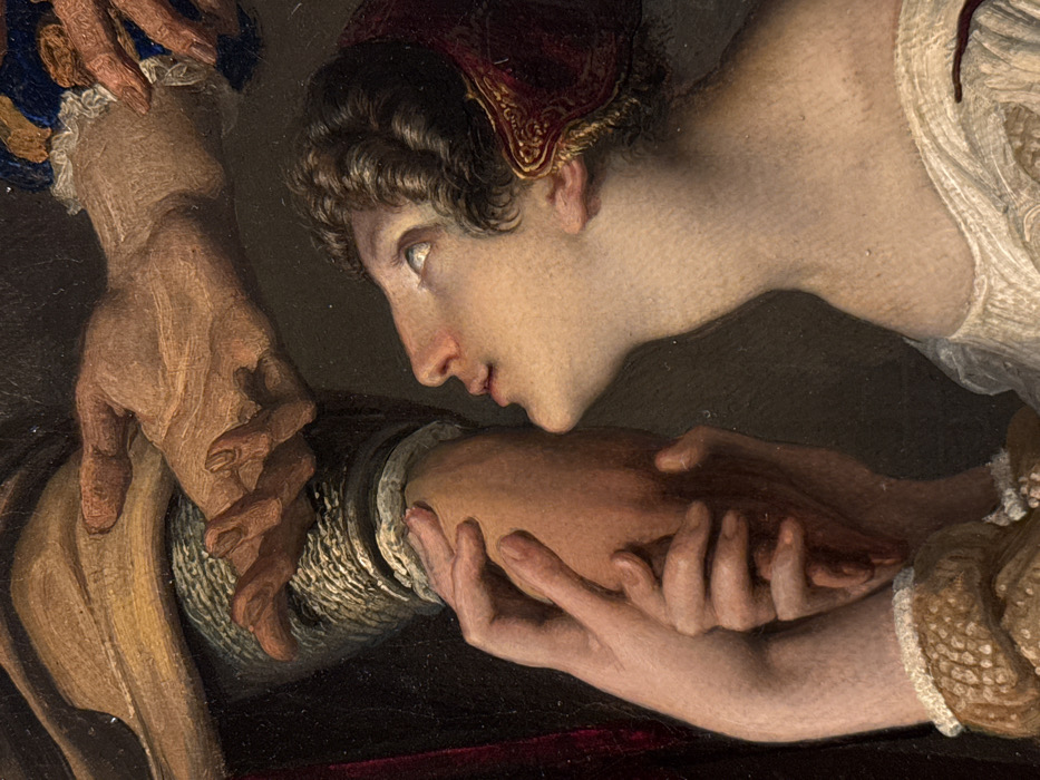 This is a detailed close-up of an oil painting, specifically a section of "The Kiss" (Il Bacio) by Francesco Hayez, located in the Pinacoteca di Brera in Milan, Italy. The scene depicts an intimate interaction between two figures: a woman on the right and a man on the left.
The woman is shown in profile, facing left and upward, with her mouth slightly agape and her eyes cast toward an unseen point above. She possesses fair skin, dark, curly hair visible beneath a red cap adorned with intricate gold trim, and wears a light-colored garment, possibly linen, with visible pleats around her neck and shoulder. Her slender hands are clasped around the forearm of the man.
The man's robust forearm is visible on the left, encased in a textured sleeve that appears woven or quilted, possibly representing chainmail. Above this, a larger, darker-skinned hand belonging to the man descends, its fingers gently curled, with a ruffled cuff in blue and yellow fabric visible at the wrist. This hand is positioned above the woman's head. The background is dark and featureless, emphasizing the figures.