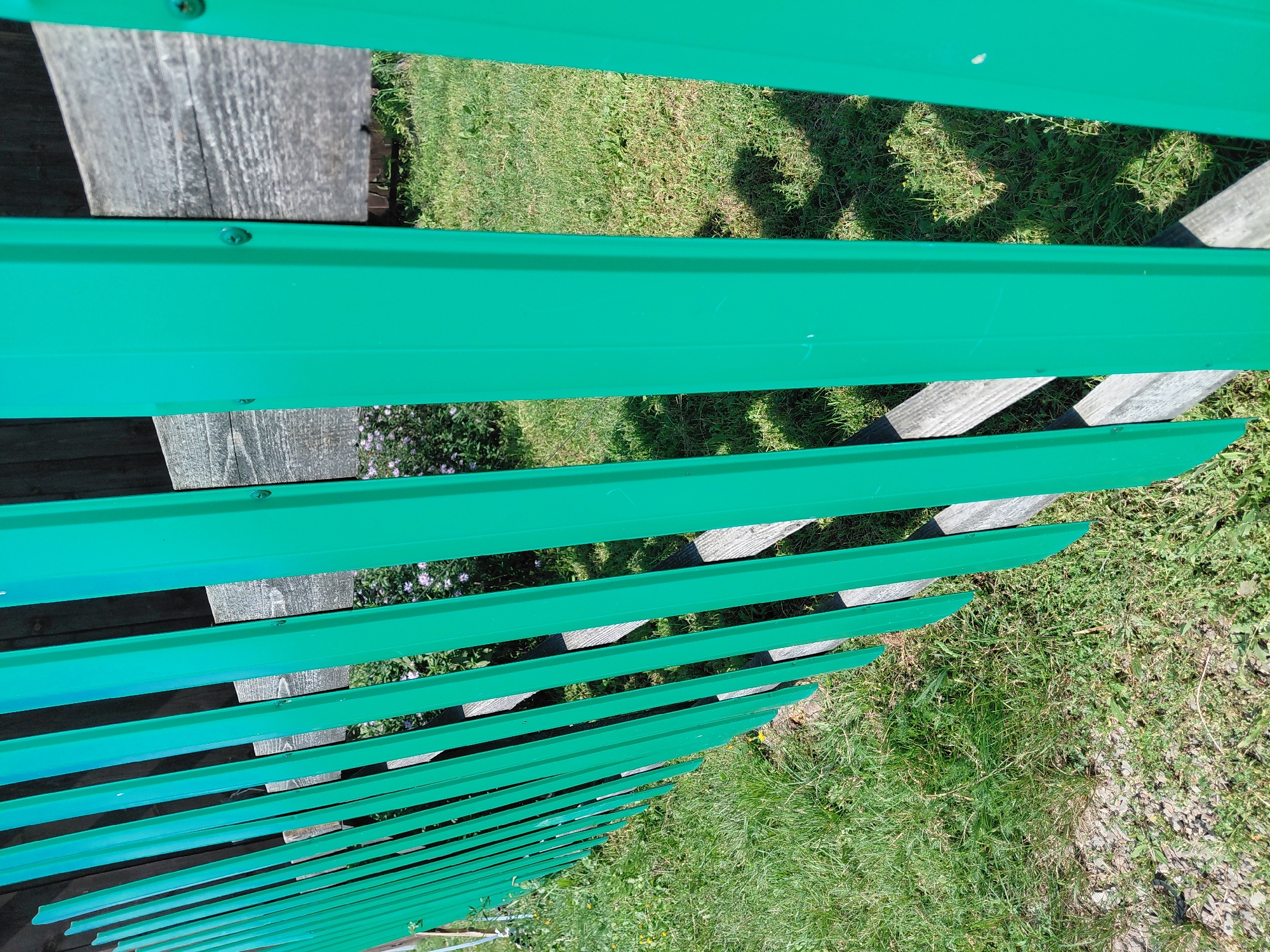 This image shows a close-up view of a green metal fence with wooden slats behind it. The fence is made of vertical corrugated metal sheets. The ground in front of the fence is covered in green grass and some gravel. In the background, there are some small purple flowers. The scene appears to be outdoors, likely in a rural or suburban setting. There are no people visible in the image, and no specific actions or interactions are depicted. The location in Malinovka, Russia is not discernible from the image itself.