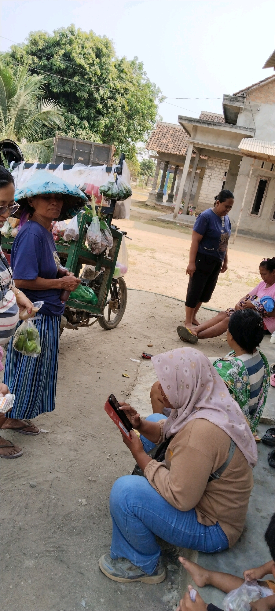 The image depicts an outdoor scene in a village setting, likely in Tanjung Ratu Ilir, Indonesia, characterized by dirt pathways and modest residential structures. In the foreground, a woman wearing a blue t-shirt and striped skirt stands beside a green mobile cart laden with goods, possibly produce, wrapped in plastic bags. She wears a wide-brimmed hat. To her left, another woman, partially visible, holds a bag of what appears to be green peppers. In the mid-ground, three women are seated. One woman, wearing a light pink headscarf and a tan shirt, is focused on a red tablet device. Another woman in a pink top sits with a baby in her lap. A third woman in a green and striped top is also seated. A man, partially visible, is also present, his legs showing. In the background, a simple house construction is visible, along with trees, suggesting a rural or semi-rural environment. The overall impression is of a casual community gathering or a marketplace interaction.