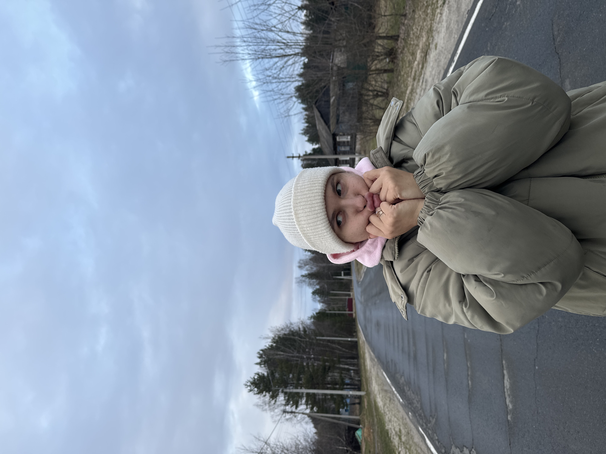 A person wearing a white beanie and light green puffer jacket is standing on an asphalt road, holding their hands to their face. The person also appears to be wearing a pink hoodie under the jacket. In the background, there is a cloudy sky, trees, a power line and a house. The location is potentially in the city of Красный Маяк, Russia.