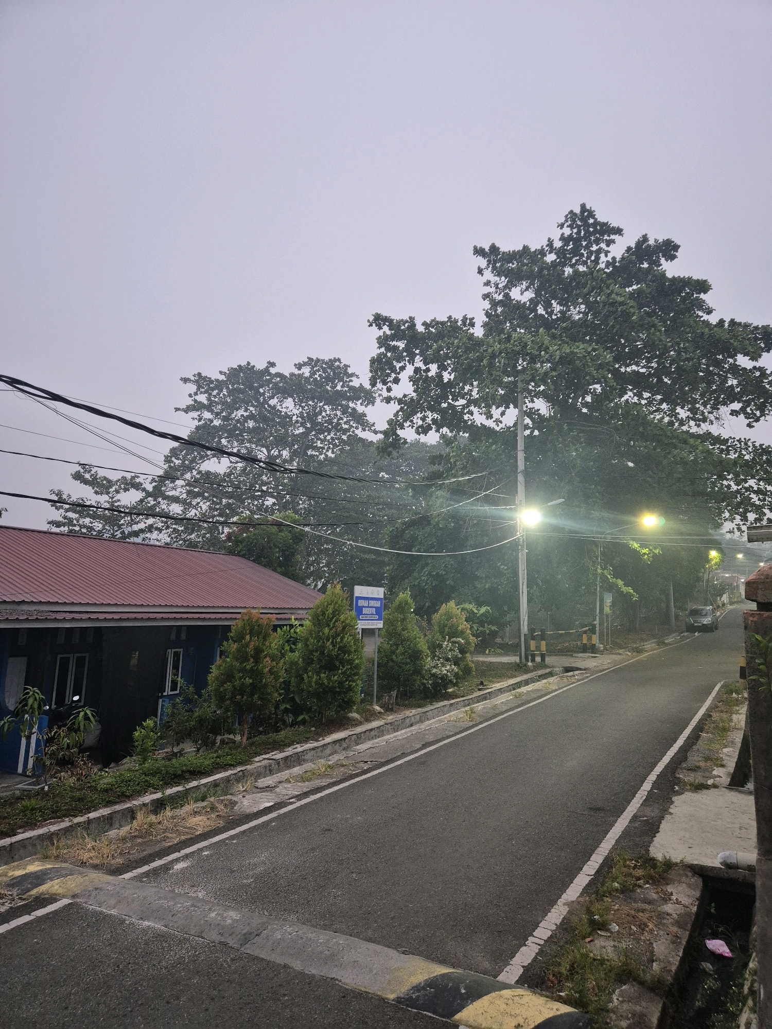 The image depicts a street scene in Bontang, Indonesia, under an overcast sky. A residential building with a red corrugated roof and blue walls is visible on the left, with power lines overhead. Adjacent to the house are several small evergreen trees and a sign that reads "RUMAH SINGGAH DINSOSPOL." A paved road stretches into the distance, illuminated by streetlights. A dark-colored car is parked on the right side of the road, near some trees. The environment appears to be a residential area with a mix of vegetation and man-made structures.