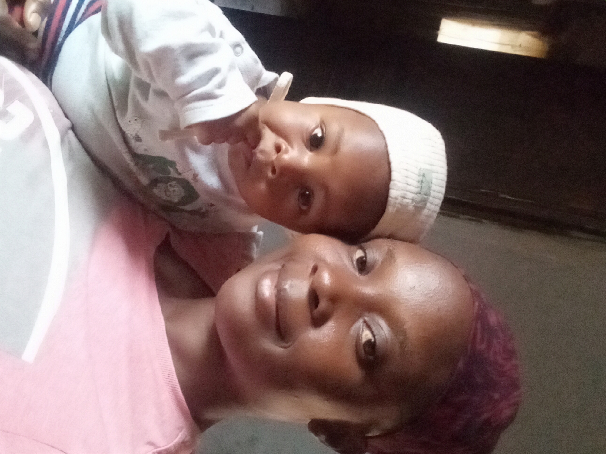 A selfie captures a woman and a baby. The woman is positioned at the bottom of the frame, and the baby is nestled above her, both facing the camera. The woman has dark skin, short red-tinged hair, and is wearing a light pink shirt. The baby has dark skin and is wearing a white knit hat and a white long-sleeved shirt. The background is dimly lit and appears to be the interior of a building. The location is Sabon Tasha, Nigeria.