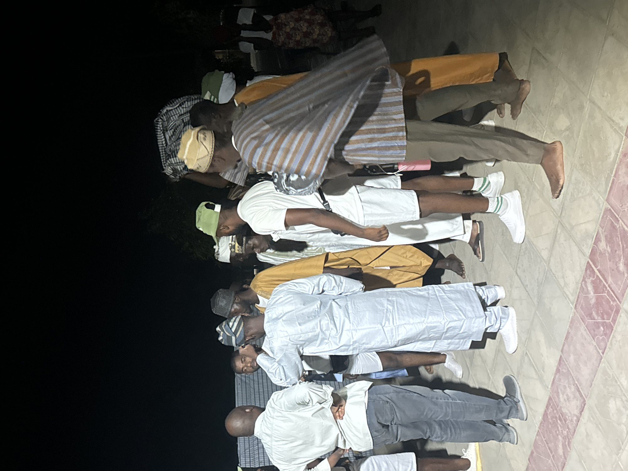 This media file captures a group of approximately ten men standing in a line or formation, appearing to be engaged in a communal activity. The scene is set outdoors or in a semi-enclosed space, likely at night, indicated by the dark background and the reliance on artificial lighting which casts some shadows. The ground beneath them is a tiled surface with a geometric pattern of light and reddish-brown squares.
The men exhibit a range of attire, blending traditional and contemporary styles. Several individuals are dressed in long white or light-colored robes, commonly known as kaftans or agbada, often paired with matching caps or kufis. Other garments include striped shirts or robes, t-shirts, and trousers or jeans. Some men are barefoot, while others wear white sneakers or sandals. Their postures are generally upright and attentive, with many looking forward or slightly downward.
The overall impression is that of a gathering, possibly a religious assembly or community event, common in places like Maiduguri, Nigeria, where such attire and group formations are often seen. No discernible text is visible within the image.