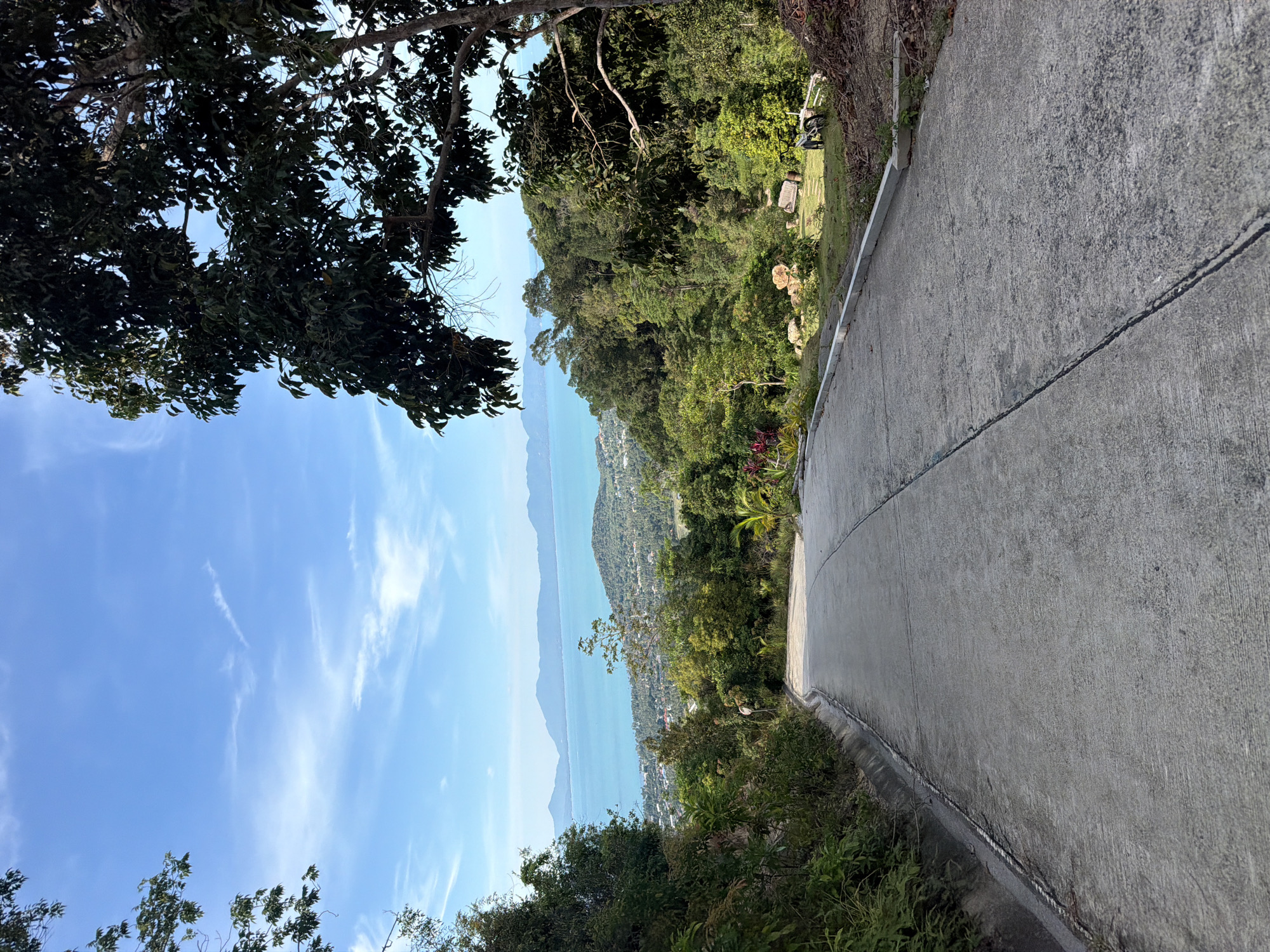 A steeply sloping concrete road descends from the foreground towards a scenic vista of turquoise water and distant islands under a bright blue sky with wispy clouds. Lush green vegetation lines both sides of the road, with dense trees dominating the right side of the frame. In the middle ground, a town with scattered buildings nestled amongst rolling hills can be seen. The background is dominated by a serene seascape and the silhouettes of mountains on the horizon. On the right side of the road, a patch of grass with a wooden bench and a bicycle is visible. The overall scene suggests a beautiful, tranquil, and elevated location, likely a viewpoint overlooking the coast of Baan Tai, Thailand.