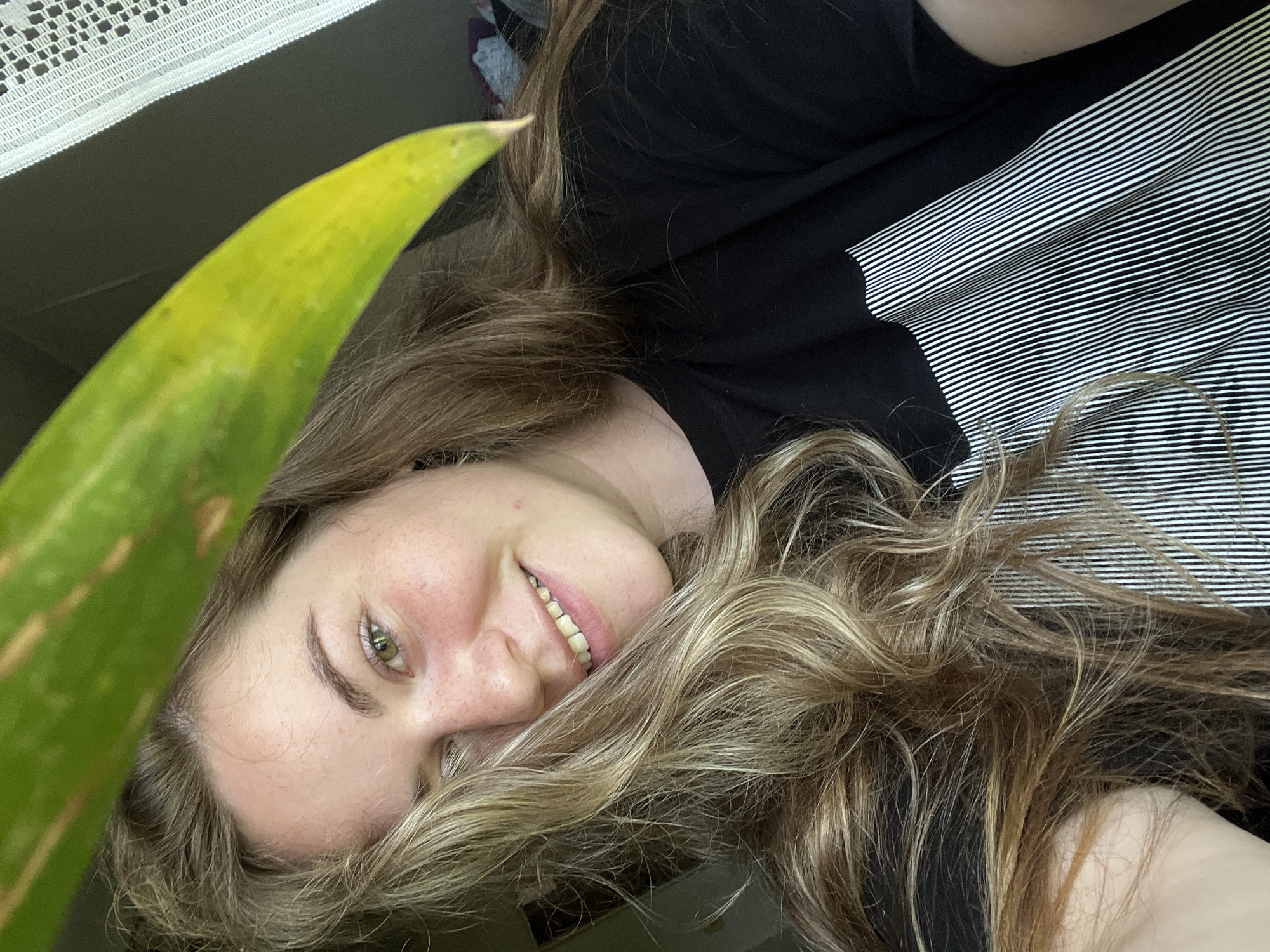 A woman with light-brown hair and green eyes is smiling and looking at the camera. She is wearing a black t-shirt with white stripes on the shoulder area. A long green-yellow leaf partially obscures her face. The background appears to be a light-colored curtain.