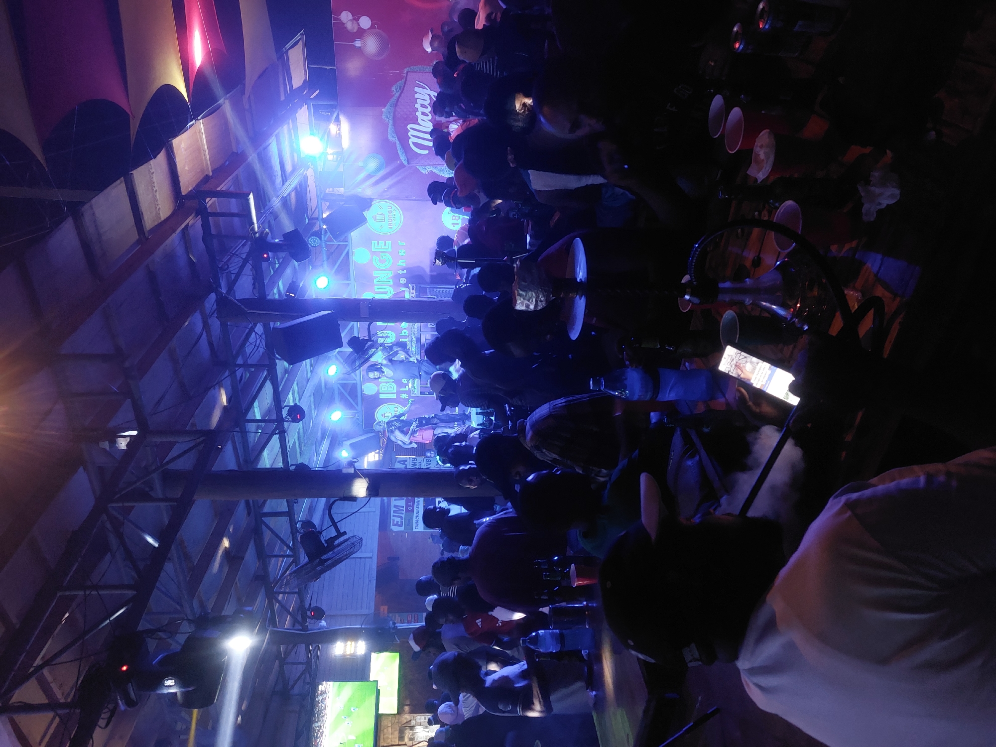 The image depicts a crowded indoor bar scene in Dar es-Salaam, Tanzania. Numerous people are gathered, facing a stage with blue stage lighting, a "Merry IBUNGU Lounge AD.." sign. Some people are seated at tables with drinks, and there's a hookah setup in the foreground with smoke present. There are TV screens visible on the left showing sports, and the establishment appears to have an open ceiling with beams and hanging lights.