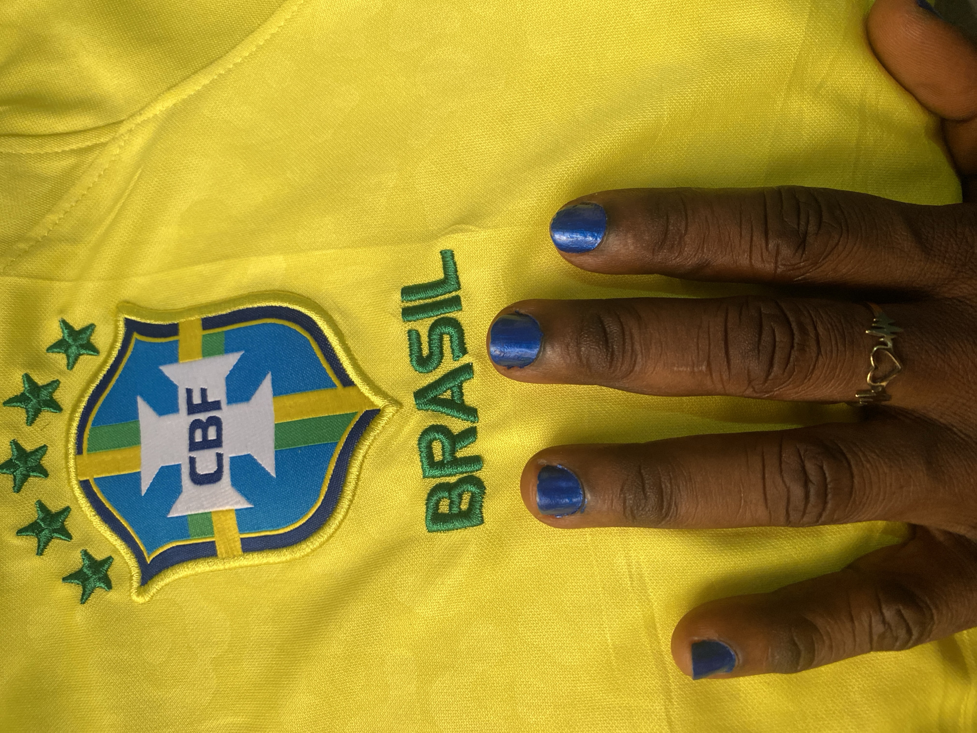 A close-up photograph shows a portion of a yellow Brazil national football team jersey with the Brazilian Football Confederation (CBF) crest visible on the left. The crest features a blue and yellow shield with the acronym "CBF" in white, surrounded by five green stars. Next to the crest, the word "BRASIL" is embroidered in green. A dark-skinned hand with blue nail polish rests on the jersey. The ring finger of the hand has a gold-colored ring with a heart design. The background is the yellow fabric of the jersey. There is no evidence in the image to determine a specific location in Lokoja, Nigeria or any other location.