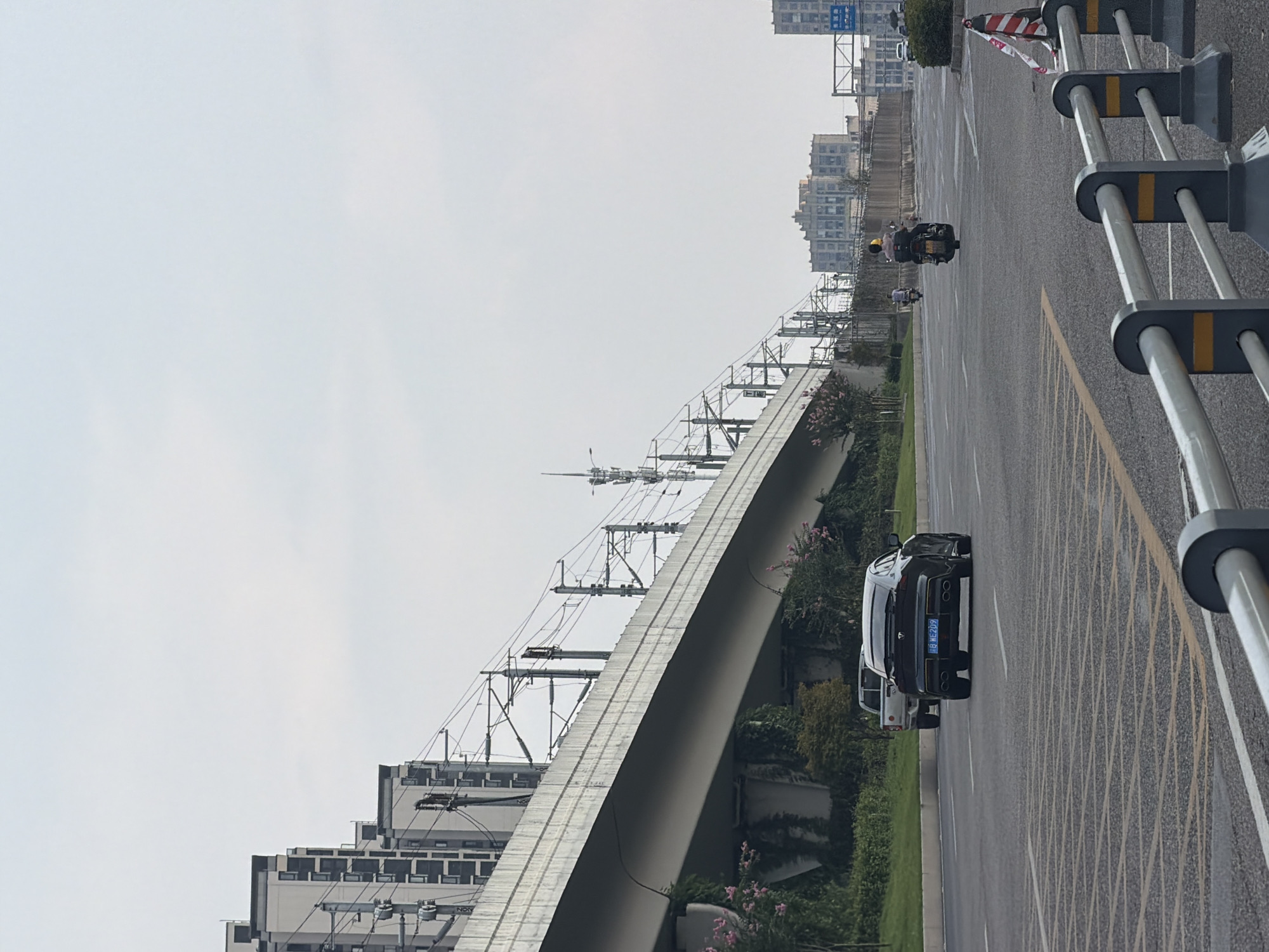 An outdoor, daytime, low-angle, vertical, medium shot shows a road in the city of Jiangjin, China, with a highway overpass running diagonally from the left middle ground to the right middle ground. The sky is overcast with a pale blue-gray hue. In the foreground, the asphalt road surface is marked with yellow lines and a grid pattern. A silver railing with dark gray and yellow supports runs along the bottom of the frame.
On the road, moving away from the viewer, is a black four-door sedan with tinted windows and quad exhaust pipes. Its license plate reads "B WE209." To the left of the sedan, a silver pickup truck is also moving away from the viewer. Further down the road, two people on motorcycles are also moving away from the viewer, one with a yellow helmet.
Beneath the highway overpass, lush green vegetation, including trees and flowering bushes with pink blossoms, grows. On the left side of the image, a row of modern apartment buildings with light gray facades and dark roofs rises into the sky. The overpass itself is a wide, light gray concrete structure supported by concrete pillars. Numerous electrical wires and utility poles are visible along the overpass, indicating it is likely a transportation corridor. In the background, more buildings are visible, some appearing older and more industrial, while others are more modern high-rises.
The overall scene depicts a bustling urban environment with infrastructure for transportation and residential living, under a somewhat muted sky.