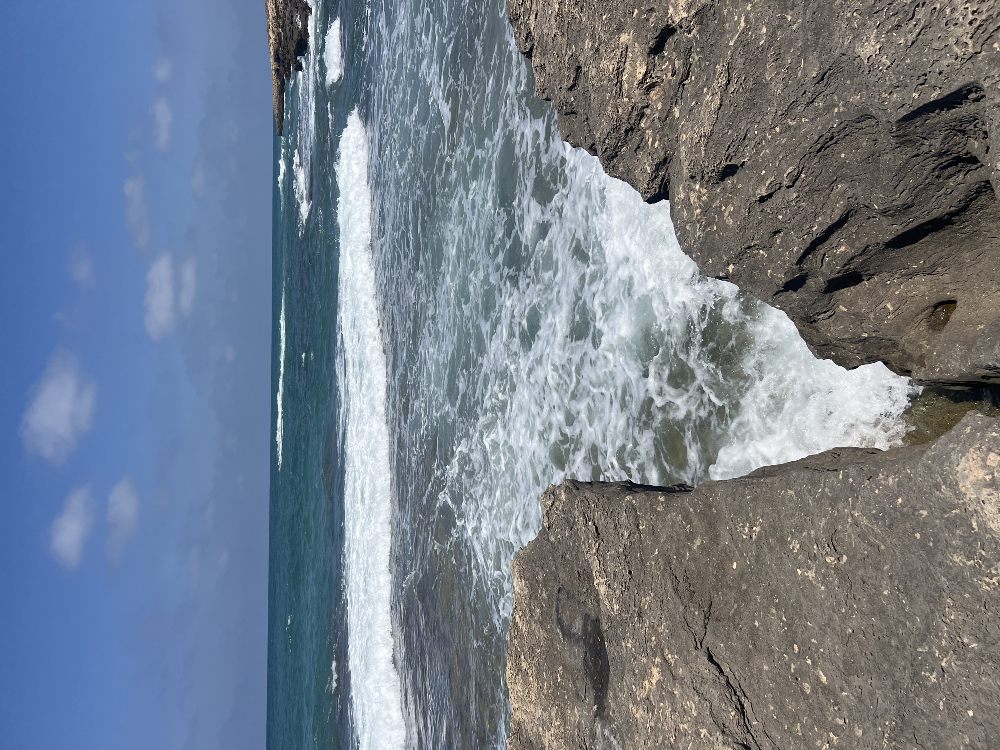 The image shows a rocky coastline with waves crashing against the shore. The water is a deep blue-green color, and the sky is a clear blue with a few scattered clouds. In the foreground, there are large, gray rocks that appear to be weathered and worn by the sea. The waves are white and foamy as they break against the rocks. The scene is peaceful and serene, with the sound of the waves being the only audible element. This coastline is part of the city of Hof HaCarmel Regional Council, Israel. There are no people or animals visible in the image. The overall impression is one of natural beauty and tranquility.