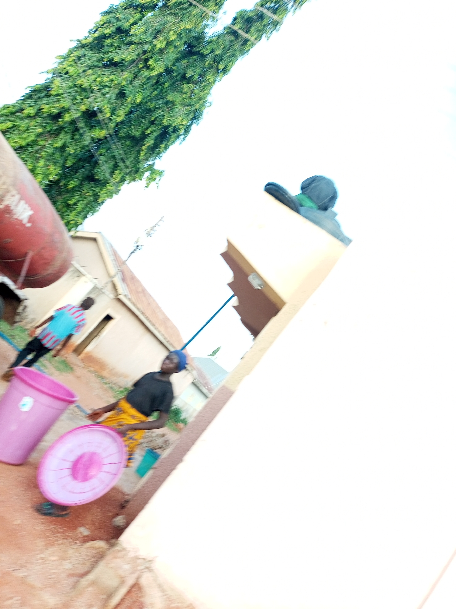 A low-angle, wide shot captures an outdoor scene in what appears to be a residential area in Tempure, Nigeria. The image is heavily tilted, giving a dynamic perspective. In the foreground, to the left, is a large, bright pink plastic tub. Next to it, and slightly further back, is a flat, pink plastic lid. A person wearing a black t-shirt and a yellow and patterned skirt is visible in the middle ground, to the right of the tubs, with their back partially turned towards the camera. They appear to be holding or interacting with the pink lid. Further to the left in the middle ground, another person wearing a blue and red striped shirt is walking away from the camera. Behind them, a large, red, cylindrical tank, possibly a water tank, is partially visible on the far left. The background consists of light-colored buildings with corrugated metal roofs and a tall, lush green tree on the left side of the frame. The sky is bright white, suggesting it is daytime. The ground appears to be a mixture of dirt and some scattered debris. There is a blue pipe running diagonally across the scene from the right towards the buildings. A dark object, possibly a hat or bag, rests on a light-colored surface on the right side of the image.