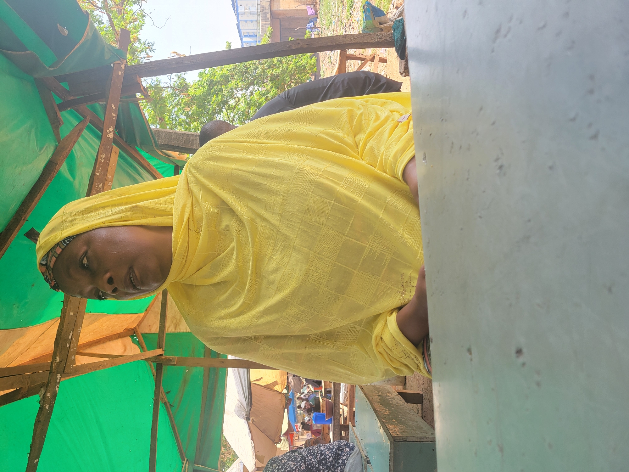 The image, originally rotated, captures an outdoor scene, likely a bustling market or roadside stall in Abuja, Nigeria, illuminated by bright daylight under a clear sky.
The primary subject is a woman positioned in the left foreground, looking slightly downwards and to her left with a neutral expression. She wears a vibrant yellow headscarf (hijab) and a matching long-sleeved garment, which features a distinct, textured checkered pattern. Her right arm and hand are visible towards the right side of her body.
She is situated within a makeshift structure, characterized by a canopy of green tarpaulin stretched over rough wooden poles and beams, suggesting a temporary shelter or stall. To the immediate right, a large, light grey or blueish-grey vertical surface partially frames the scene, possibly a wall, partition, or a large object.
In the background, other individuals are visible. One person wearing a black garment is partially obscured by the foreground woman. Further back, more people and various informal structures can be seen, along with what appears to be a blue plastic container. Trees and sparse vegetation are visible against the bright sky, indicating an open-air environment.
A visible detail in the distance, particularly clear in one of the cropped sections, is text on a building sign that reads "UNION BANK", suggesting the presence of commercial establishments nearby. The overall impression is a common, everyday scene of life and commerce in an urban or semi-urban setting. The woman appears to be either observing her surroundings or resting, but no specific activity is explicitly shown.