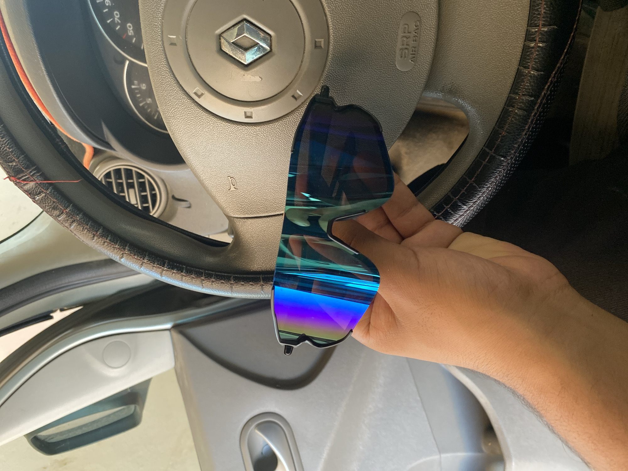 A person's hand holds a pair of blue and purple mirrored sunglasses in front of a car's steering wheel. The car's interior is visible, including the dashboard and a door panel. The scene is shot from a close-up perspective, focusing on the sunglasses and the hand. The lighting is natural, casting shadows on the objects. The background suggests an indoor setting, possibly a garage or a parking lot. There is no specific information to determine the location within the city of Aziz, Algeria.