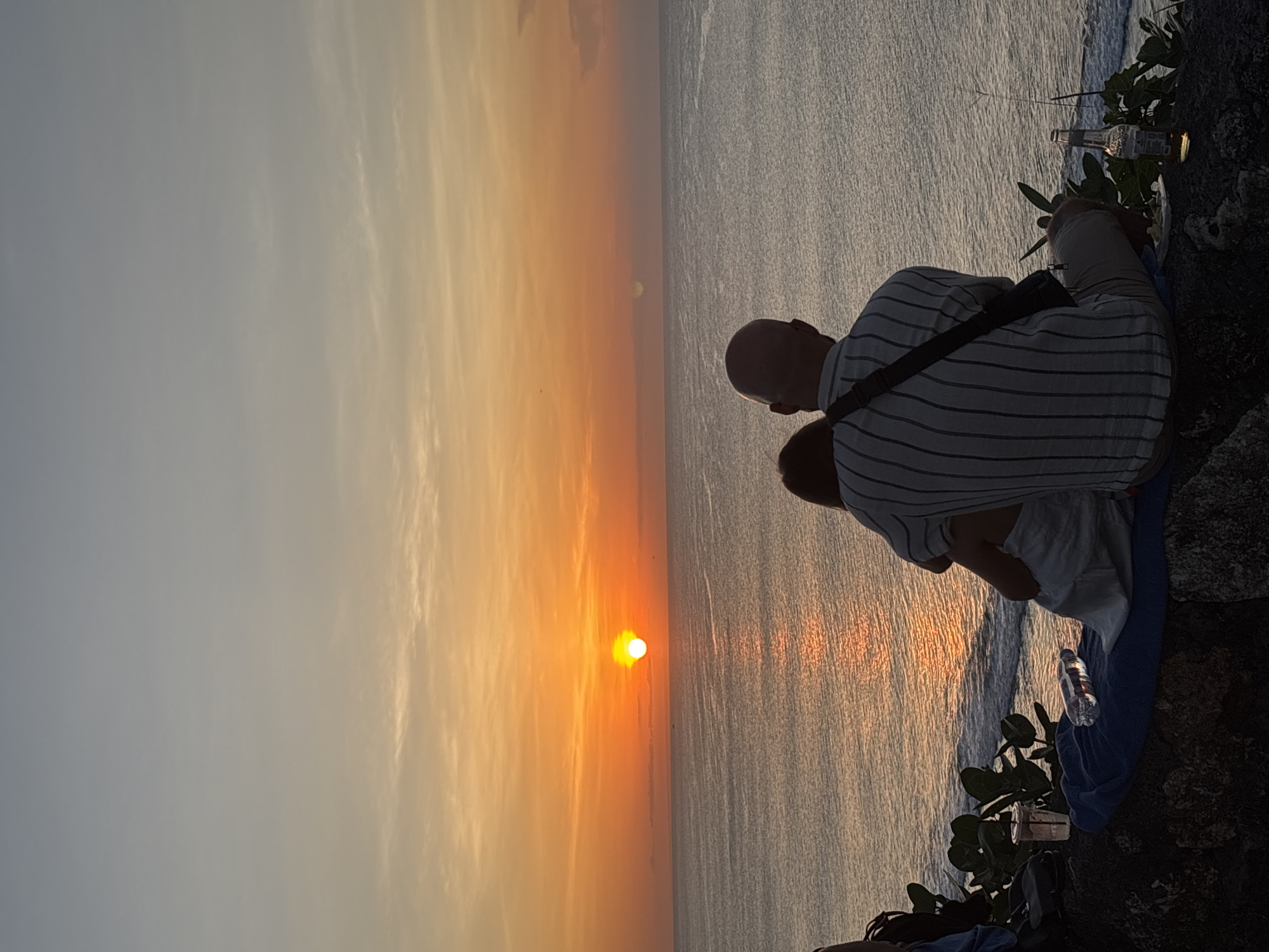 A scenic shot taken near Uluwatu Temple in Kuta Selatan, Indonesia, captures a father and child enjoying a sunset view over the ocean. The scene features a bald man wearing a striped shirt and dark-colored bag, standing beside a child. The backdrop showcases a brilliant sunset. Also visible are a water bottle, a beer bottle, and plant life. The photograph gives a vivid representation of a father and child sharing a quiet moment against the serene coastal landscape of Kuta Selatan.