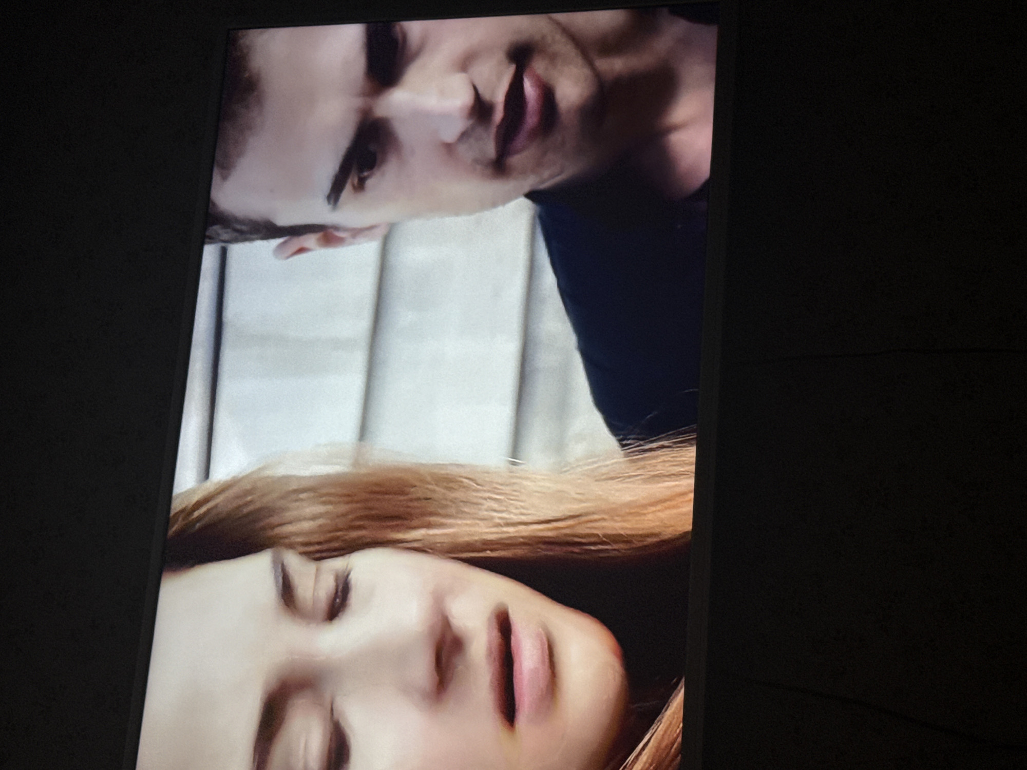 The image shows a close-up of two people, possibly from a TV screen. A man with dark hair and light skin is positioned at the top, looking down. Below him is a woman with long, reddish-brown hair, her eyes closed. They are in front of what appears to be a light-colored, possibly textured wall or surface. No location in the city of Mytishchi, Russia is discernible from the image.