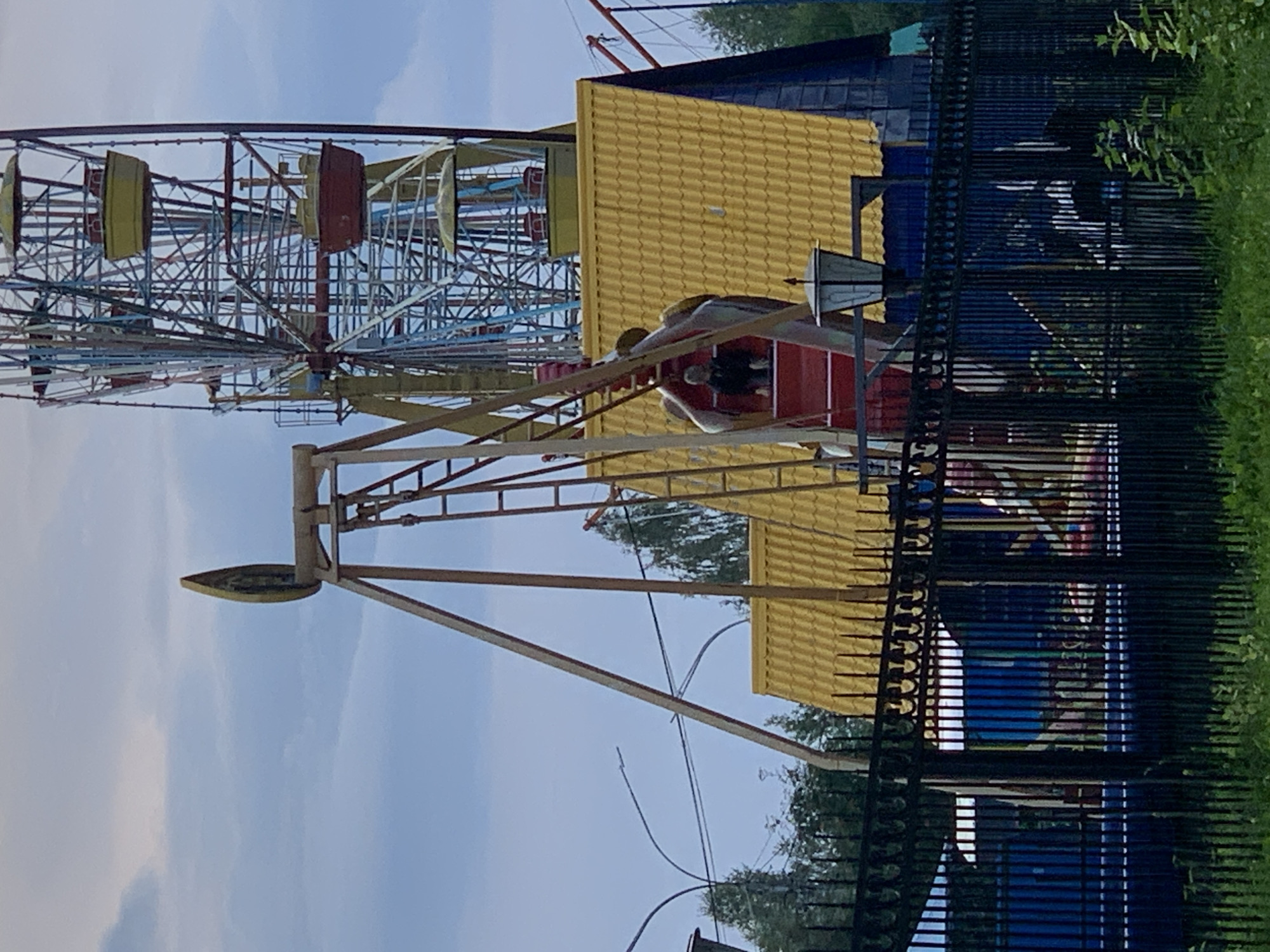 An amusement park scene is depicted in Prokopyevsk, Russia, under a cloudy sky. A Ferris wheel, painted in red, yellow, and blue, dominates the background. In the foreground, a swing ride with a red seat and yellow accents is visible. A single individual is seated in the red swing. The amusement park is enclosed by a black metal fence. Beyond the fence, a yellow-roofed building with blue walls is situated. Trees are visible behind the structures, and grass and foliage line the bottom of the frame.