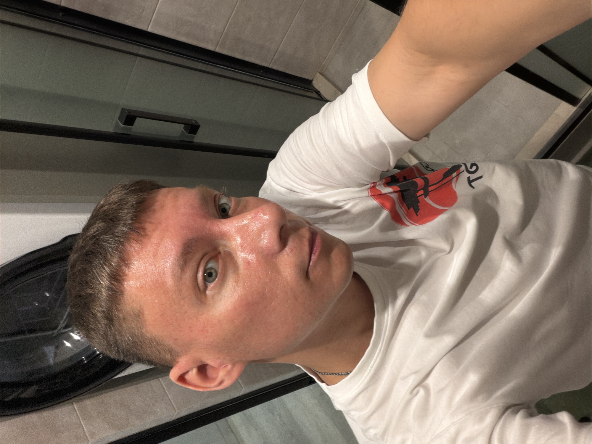 A Caucasian male with short brown hair and blue eyes is taking a selfie in what appears to be a bathroom. He is wearing a white t-shirt with a graphic design on the front. The background shows tiled walls, a glass shower door with a black handle, and what looks like a washing machine. The man's face is slightly flushed and appears sweaty, suggesting he may have just exercised or is in a warm environment. The overall lighting is dim. The image was taken in Moscow, Russia.