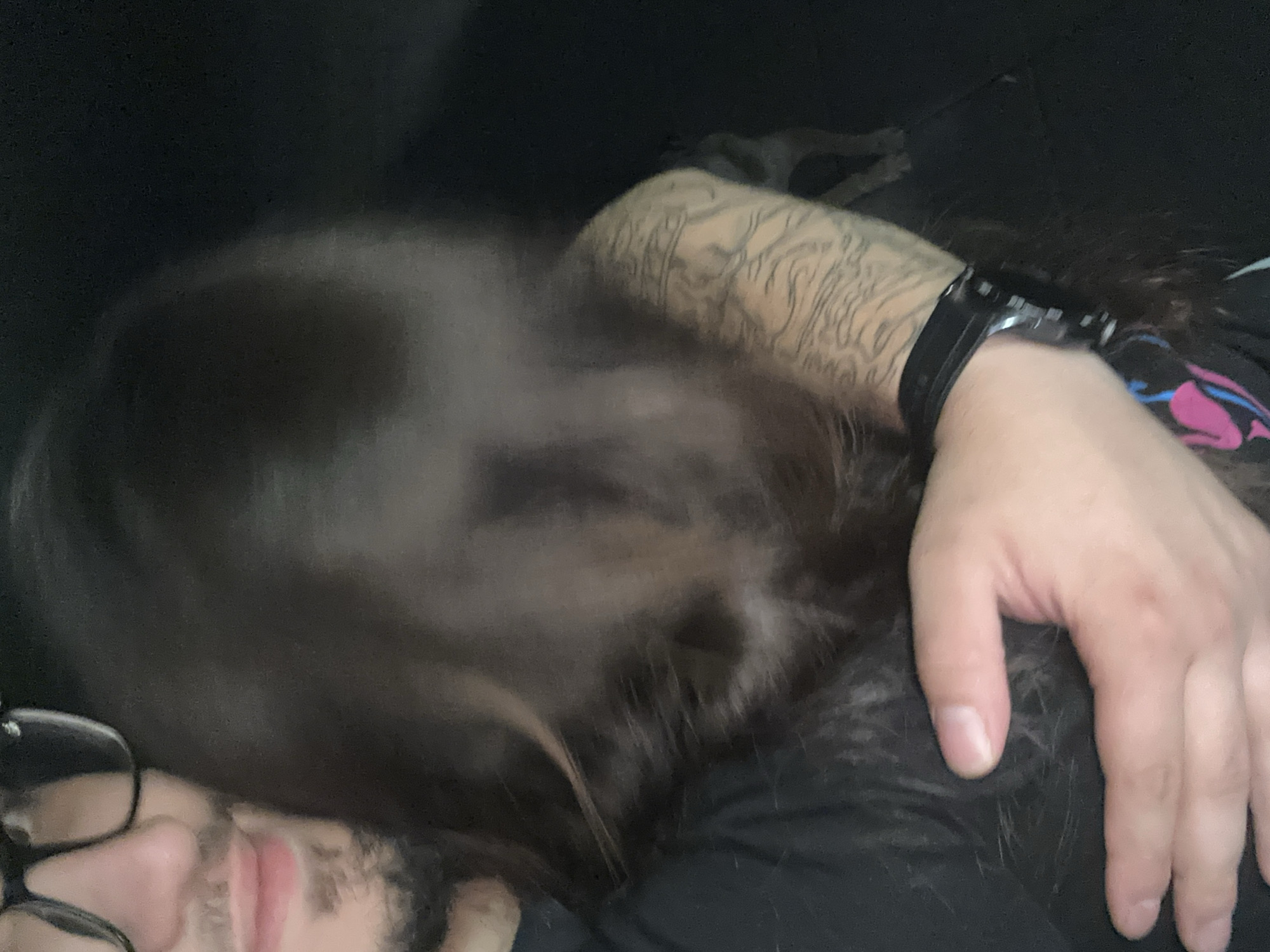 The image depicts a close-up, intimate scene taking place indoors, with a focus on a person resting. In the bottom left, part of a person's face is visible, showing glasses, a nose, a slightly open mouth, and a beard. Their expression appears relaxed, possibly asleep or dozing. Long, dark hair spreads across a significant portion of the frame, covering most of the mid-section. This hair likely belongs to the visible person or another individual they are interacting with.
The person's right arm, heavily adorned with intricate sleeve tattoos, extends from the upper left towards the center. A black smartwatch is worn on their right wrist. Their right hand rests gently on what appears to be another person's head or upper body, covered by the same long, dark hair, suggesting a moment of closeness or cuddling.
The background is dark and indistinct, hinting at a dimly lit environment, possibly a bedroom or living room. The overall ambiance is calm and peaceful, conveying a sense of rest and affection. There are no visible outdoor elements or specific location markers within the frame to confirm the Lom, Bulgaria context, nor is any text visible. The time of day appears to be evening or night due to the low lighting.