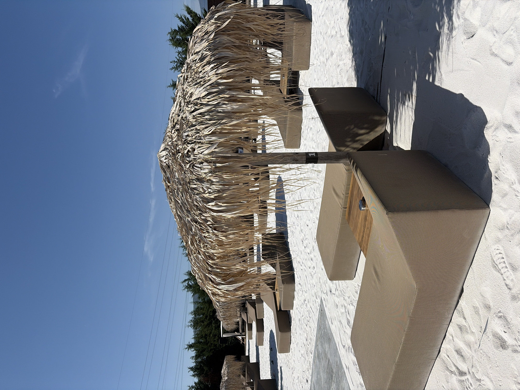 The image depicts a beach resort area with white sand and lounge chairs under a thatched umbrella. The scene is set in Kazichene, Bulgaria. There are no people visible in the image. The environment appears to be a well-maintained outdoor recreational space, likely a beach or a pool area, with rows of unoccupied lounge chairs and umbrellas. The sky is clear blue, suggesting a sunny day. The focus is on the arrangement of furniture and the natural elements of the setting.