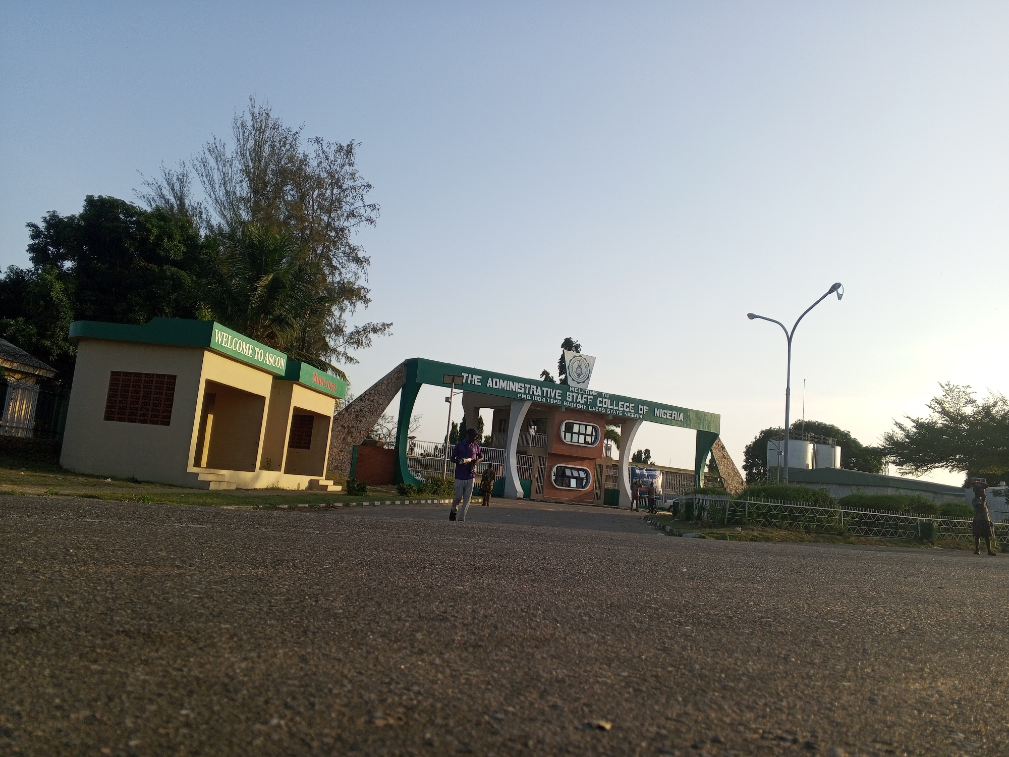 The image depicts the entrance to the Administrative Staff College of Nigeria (ASCON) in Tawpo, Badagry, Lagos State, Nigeria. A sign reads "WELCOME TO ASCON" and identifies the college. A person is seen walking through the entrance gate, and other people are visible in the background. A street light and surrounding buildings are also present. The environment appears to be outdoors on a sunny day.
