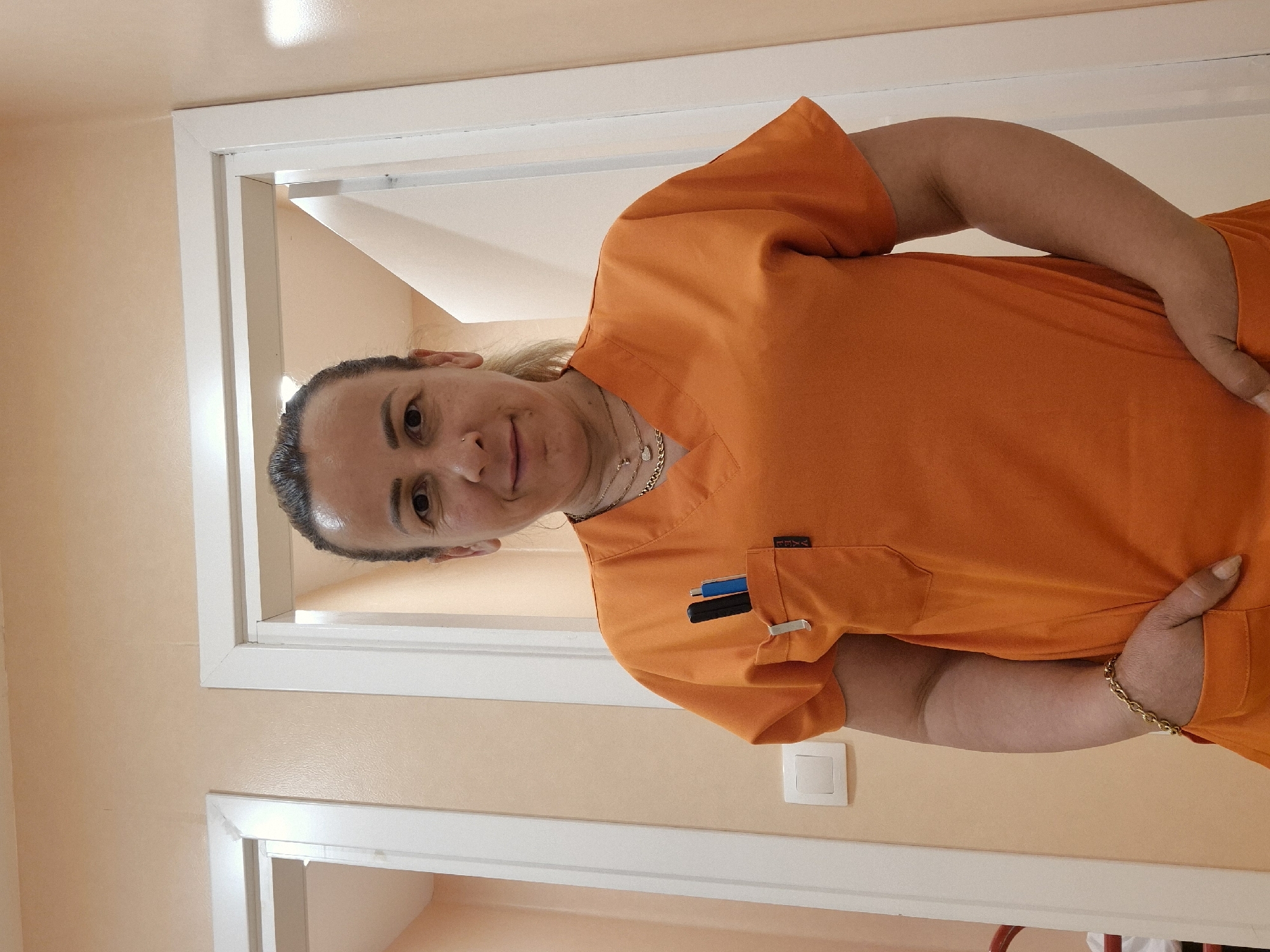 The image, originally rotated 90 degrees counter-clockwise, depicts a woman in a professional indoor setting. The woman, appearing middle-aged with pulled-back dark hair and lighter highlights, is looking directly at the camera with a calm expression. She is wearing an orange medical scrub top with a left chest pocket holding a blue pen, a black pen, and a silver clip. A small tag with the text "TRVA" is visible near the pocket. Around her neck are multiple layered necklaces, including silver and gold chains with pendants. She also has a nose piercing. On her right wrist, she wears a gold chain bracelet, and her natural, slightly long fingernails are visible on her right hand, which is placed on her hip or in a pocket. The background consists of plain light peach or beige walls and white doorframes. A white light switch or electrical outlet is visible on the wall in the lower right portion of the frame. The lighting appears to be bright indoor lighting, creating some reflections on the upper wall and her hair. The woman is standing still, posing for the photograph.