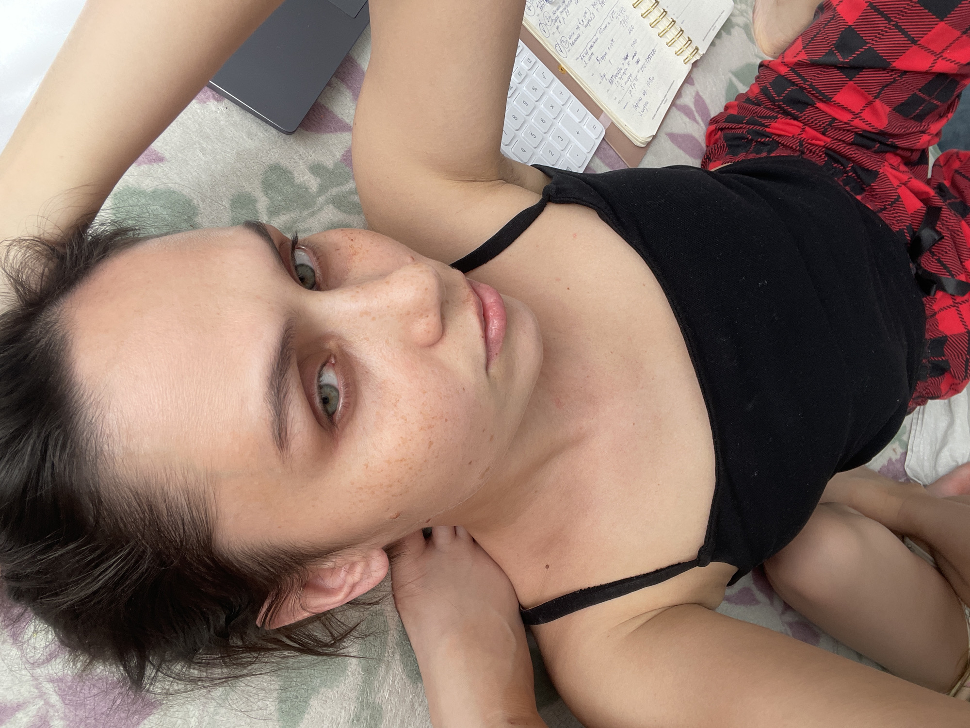 A young woman with fair skin, freckles, and light-colored eyes lies on a bed. She wears a black tank top and red plaid pajama bottoms. Her dark hair is disheveled around her head. To her left, a laptop and a notebook rest on the bed. The notebook has writing in it, and a white calculator is next to it. Her feet are near her torso. The bed is covered with a patterned sheet. The photo is taken from a high angle, looking down on the woman. There is no information about the city of Izhevsk, Russia.
