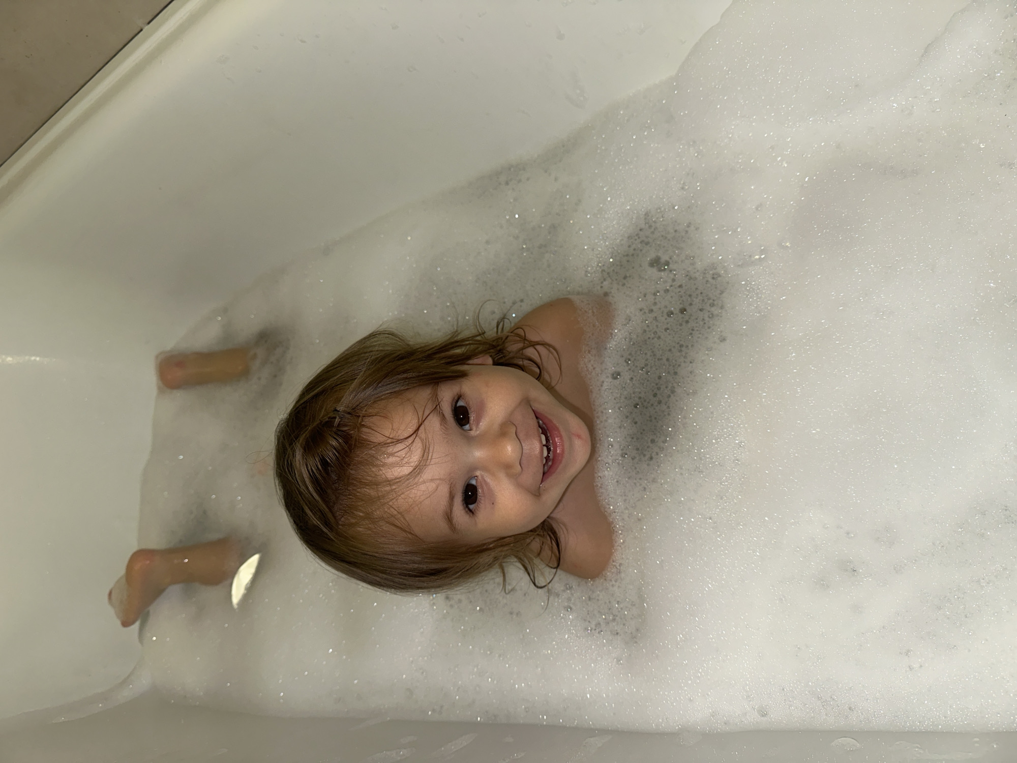 A young child is in a bathtub filled with bubbles. The child is lying on their back and smiling at the camera. Their head is tilted towards the top of the image. The child's feet are visible at the bottom left of the image. The bathtub appears to be white. The location is not specified as the city of Kharnavy, Russia.