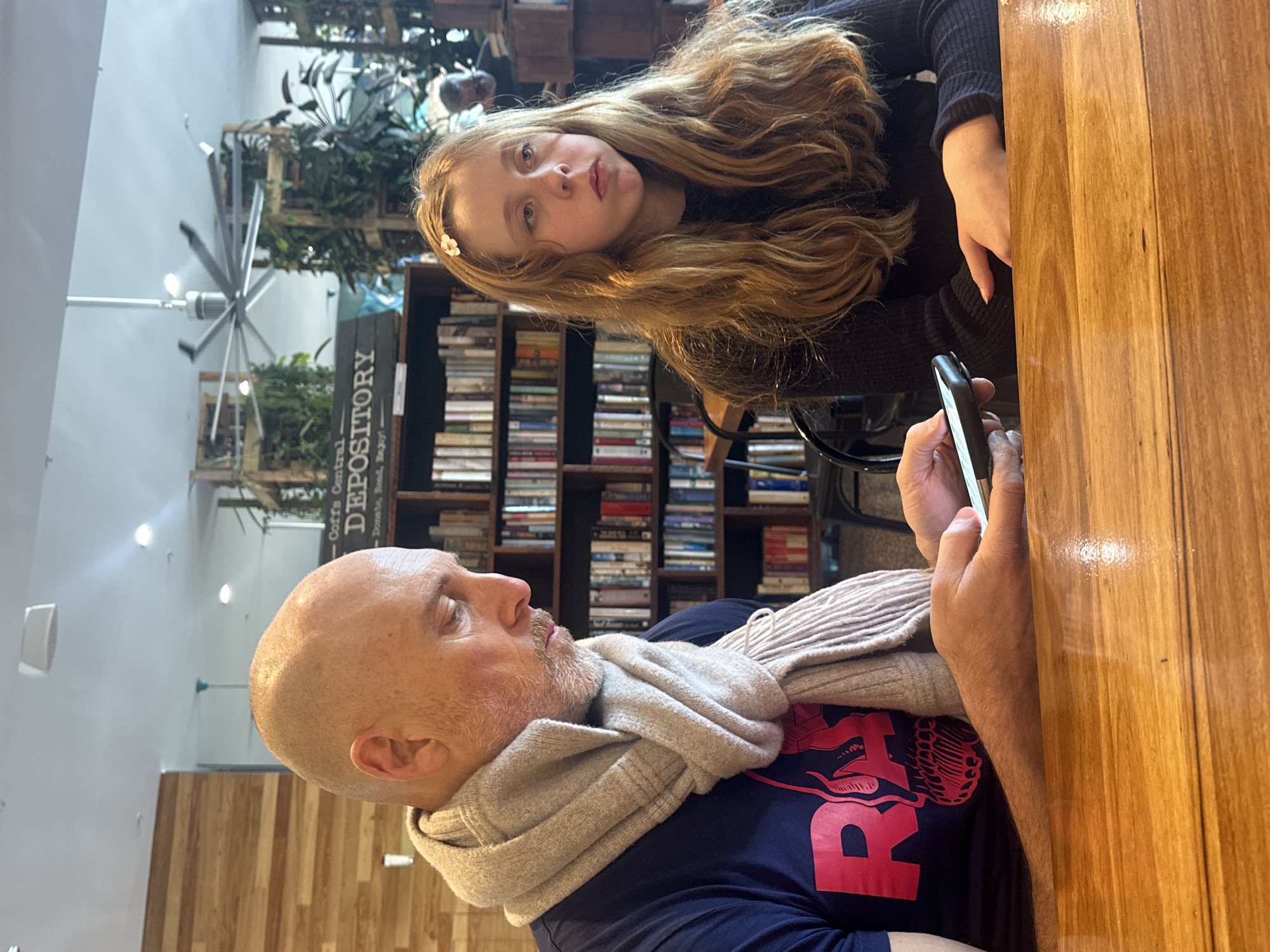 This is a medium shot of a bald man and a young girl sitting at a wooden table in what appears to be a cafe or bookstore in Coffs Harbour, Australia. The man, wearing a navy blue t-shirt with a red graphic and a light gray scarf, is looking down at his smartphone, which he is holding in his left hand. The girl, with long blonde hair and a small white flower clip, is looking to her right with her mouth slightly open. Behind them are bookshelves filled with books and decorative plants, suggesting a public space with a focus on literature and greenery. A ceiling fan is visible in the background. The overall lighting suggests an indoor environment.