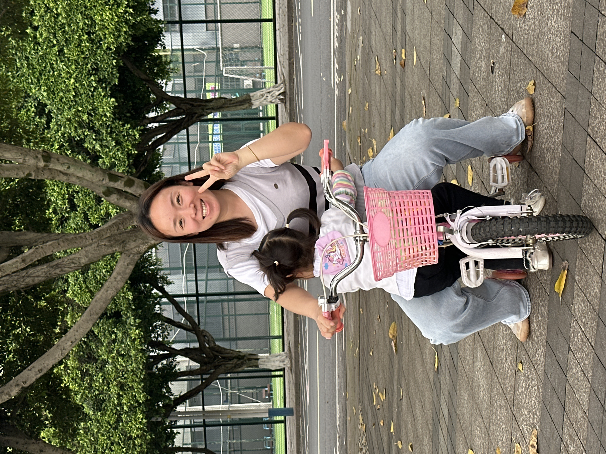 The image shows an adult woman and a young child on a bicycle on a paved area, likely a sidewalk, next to a road in Jiangjin, China. The woman is smiling and holding up a peace sign with one hand. She is wearing a white shirt and light-colored jeans. The child is sitting in front of her on the bicycle, which has a pink basket. The paved area is gray and has some fallen leaves scattered on it. Behind them is a metal fence and some trees.