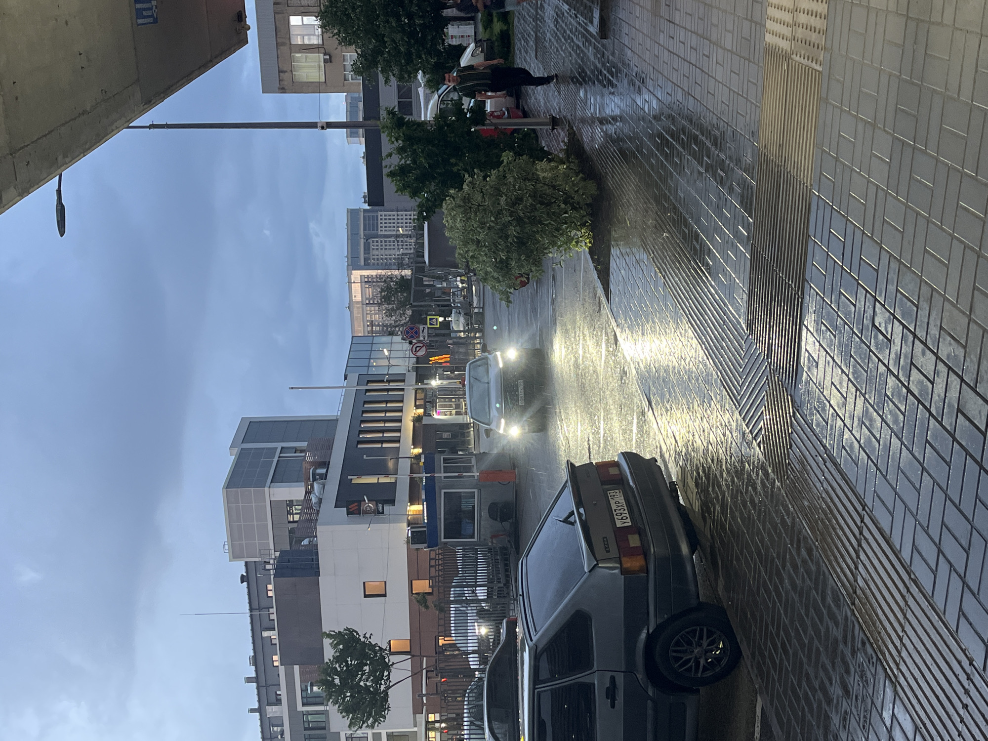 The image shows a street scene in Krasnodar, Russia, under an overcast sky, with wet pavement indicating recent rain. In the foreground, a grey hatchback car is parked, its license plate reading "Y693XP 193". To the right, a pedestrian walkway is visible with dark, reflective paving stones. Further down the street, a dark sedan is driving away, its headlights illuminated. A man in a dark green striped shirt and black pants walks away from the camera on the pedestrian walkway. In the background, modern buildings with glass facades and industrial-style architecture are present, some featuring balconies and rooftop elements. There is a small kiosk or booth with an orange barrier in front of it. Trees and shrubbery are scattered throughout the scene. The overall atmosphere is subdued due to the weather and lighting conditions.