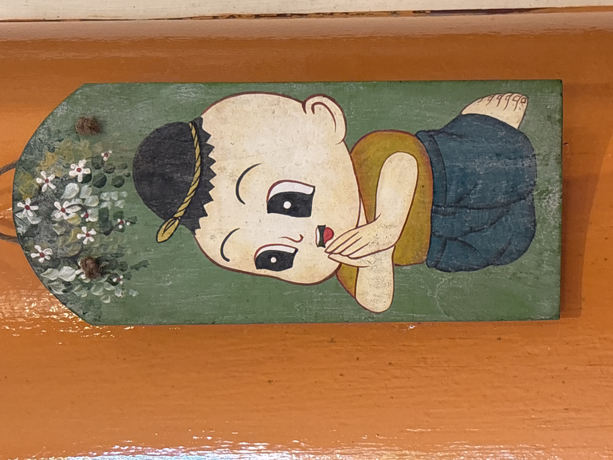 A close-up, eye-level, vertical shot captures a decorative wooden plaque painted with a cartoonish child figure. The plaque is mounted on an orange, textured surface, likely a door or wall, with a lighter colored edge to the right of the plaque. The child, depicted with a round, pale face, large black eyes with white highlights, and small red lips, is shown in a kneeling position. They wear a black cap adorned with a yellow string and a mustard-yellow shirt. The lower half of their body is covered by blue shorts. The child's hands are clasped together in front of their chest in a gesture of greeting or reverence, a common Thai tradition known as "wai." Above the child's head, the plaque is decorated with painted white flowers with red centers and green leaves, with a small, fuzzy, brown object, possibly a natural fiber or insect, attached to the artwork near the flowers. The background of the plaque is a solid green. The plaque is located in the city of Chalong, Thailand.