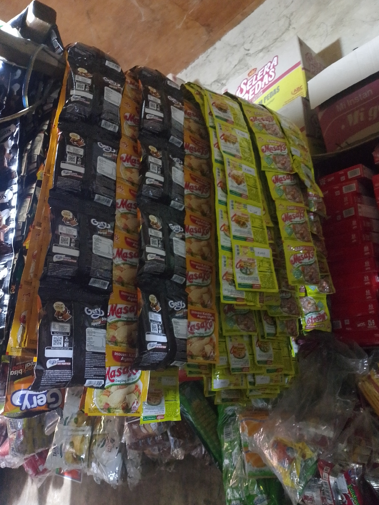The image depicts a close-up, low-angle view of a wall-mounted display of assorted packaged food products, primarily instant seasoning and noodle packets, in what appears to be a small shop or market stall. The products are hanging in rows and are largely obscured by shadows and the density of the display. Prominent brands visible include "Masako" and "Gery." Behind the hanging packets, other products are stacked, including boxes with text like "SELERA PEDAS" and "Mi Instan." The environment suggests a humble commercial setting. The lighting is dim, with the main light source appearing to be from above and to the left. The texture of the wall behind the products suggests concrete or plaster. The scene is located in Palembang, Indonesia.