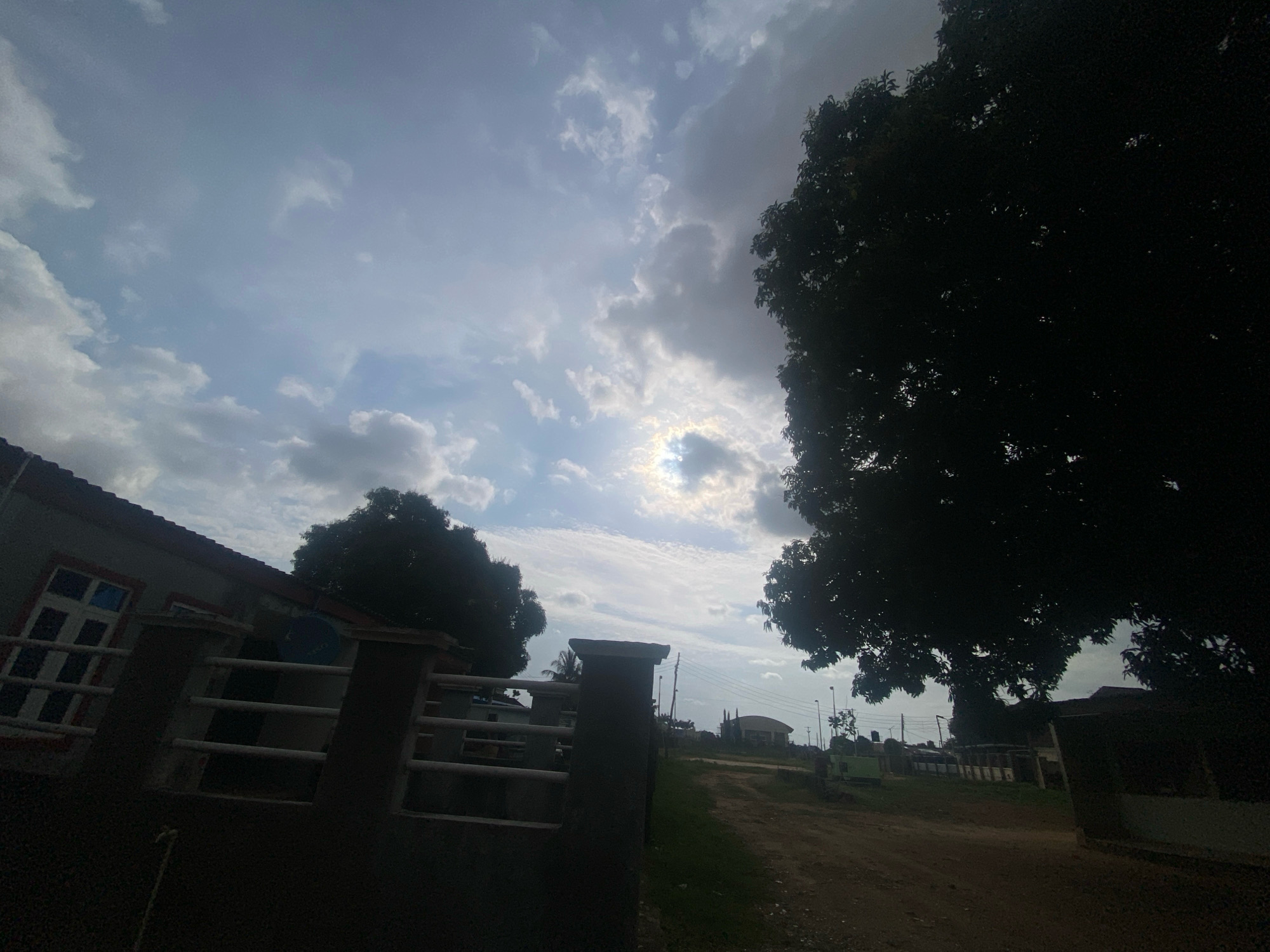 The image depicts a street scene in Barden Yana, Nigeria, under a cloudy sky. In the foreground, a dark silhouette of a large tree dominates the right side of the frame. To the left, a portion of a building with a white-railed fence is visible. The fence has horizontal bars and concrete posts. A dirt road leads away from the viewer into the distance, flanked by sparse vegetation and other structures. The sky is filled with varying cloud formations, with a bright spot suggesting the sun's presence behind the clouds, creating a diffused light. There are no people or animals clearly visible in the scene. The overall atmosphere is naturalistic and environmental, capturing a slice of everyday life in the locality.