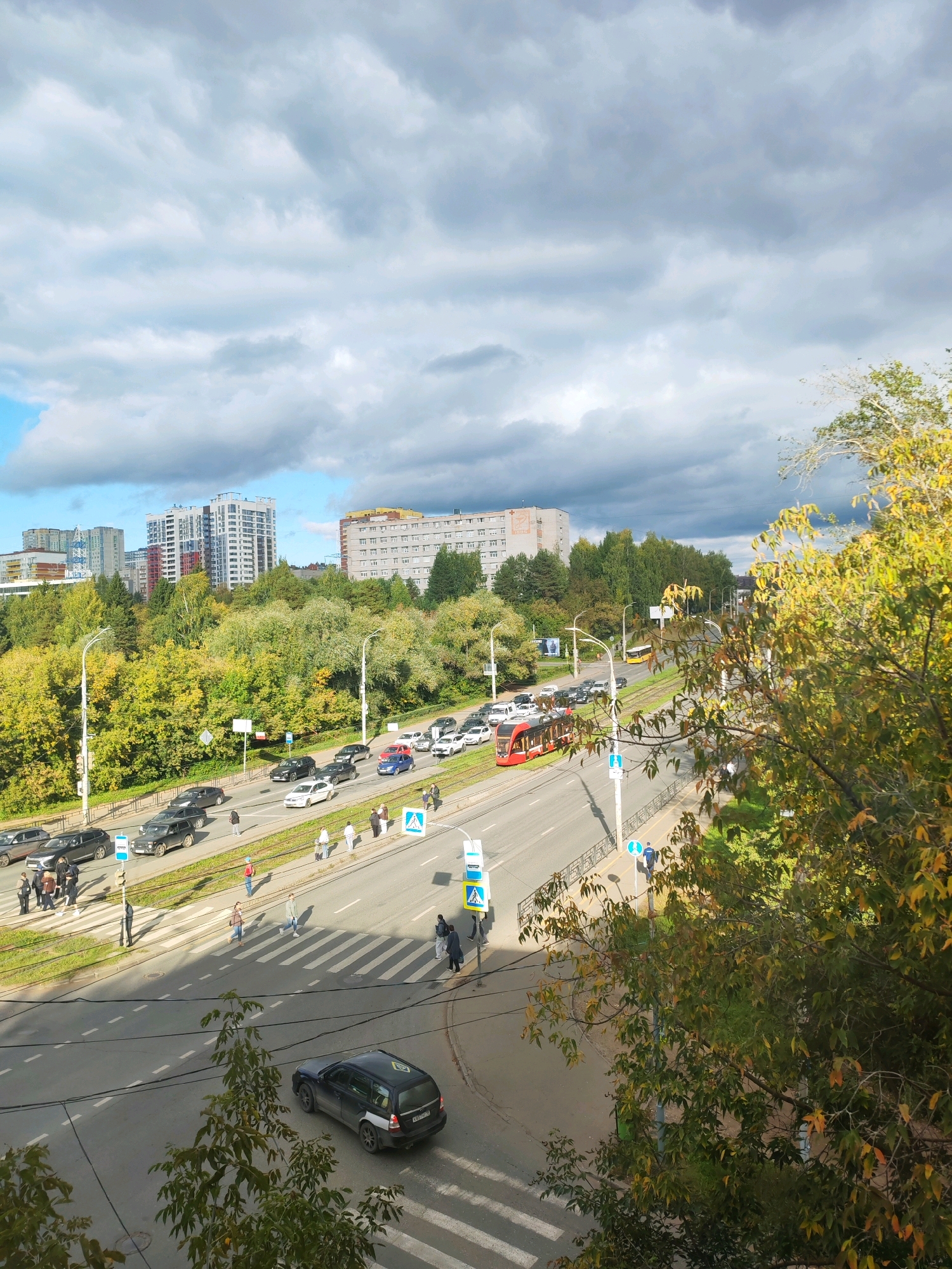 This is an aerial, eye-level, full shot of a street scene in Izhevsk, Russia, featuring a multi-lane road with moderate traffic, pedestrians, a tram, and several buildings in the background. The image captures a section of ulitsa 10 Let Vladvostoka near the intersection with ulitsa Lva Tolstogo.
On the road, a red tram is traveling from left to right. Several cars, including a blue sedan, a white sedan, and black SUVs, are also visible, with some stopped in traffic. Pedestrians are crossing the street at designated crosswalks. Some are on the sidewalks, while others are in the process of crossing.
The surrounding environment is a mix of urban and natural elements. Lush green trees with patches of yellow foliage line the road and extend towards the background, indicating an autumn season. In the distance, several multi-story residential buildings are visible, characterized by their modern architecture and varying colors. The sky is partly cloudy, with large, gray cloud formations dominating the upper portion of the frame, interspersed with patches of blue sky. Power lines crisscross the foreground, casting shadows on the road.
The overall impression is a busy urban street during daytime, with everyday activities unfolding.