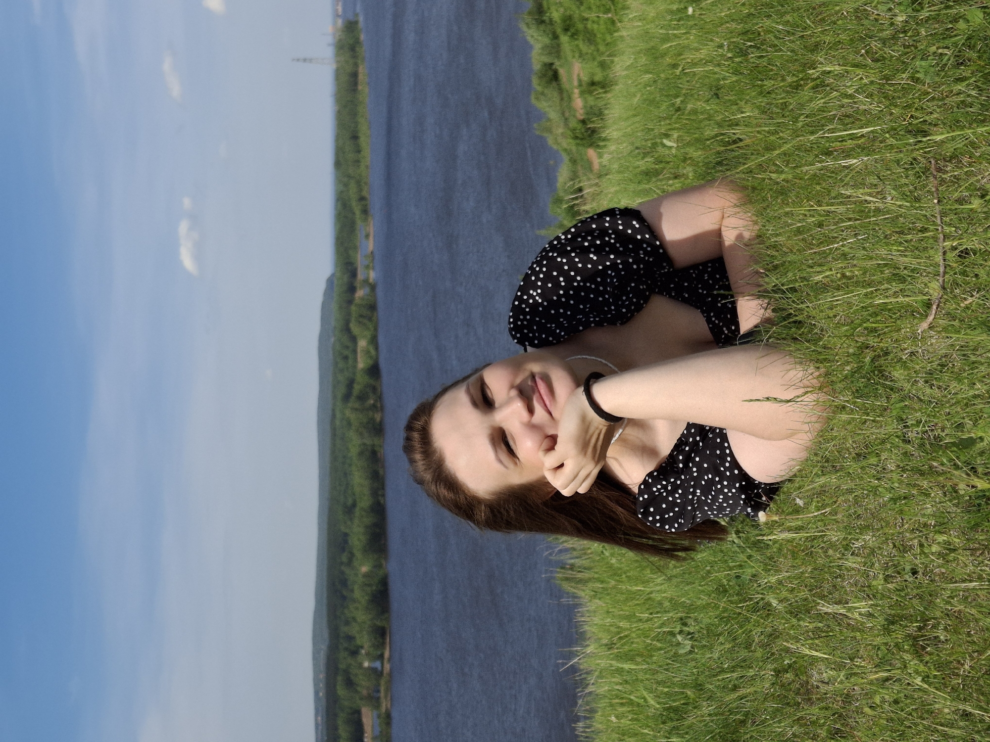 A woman is lying in a field of green grass near a body of water on a sunny day. She is wearing a black and white polka dot dress, a white necklace, and a black hair tie on her wrist. Her brown hair is spread on the grass. The body of water is a wide river with tree-lined banks. The location is Ольховка, Russia. The sky is light blue with scattered clouds.