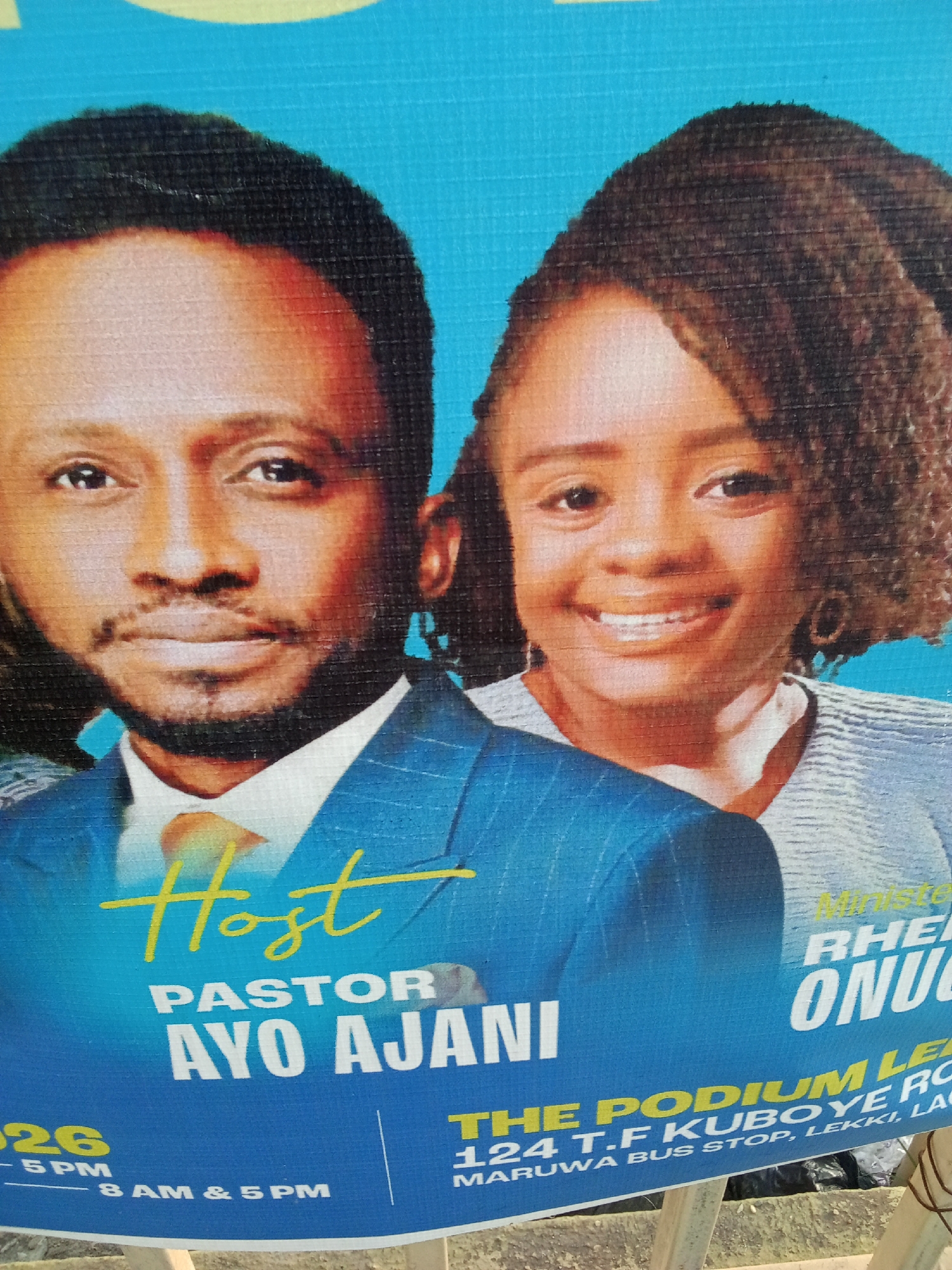 The image is a close-up of a poster advertising an event, likely a religious service or meeting. The poster features two individuals: a man and a woman. The man is dressed in a blue suit and a yellow tie and is identified as "Host Pastor Ayo Ajani". The woman is partially visible behind the man, smiling and wearing a light-colored top. The poster also contains the event details, including the location "The Podium Lekki, 124 T.F Kuboye Rd, Maruwa Bus Stop, Lekki, Lagos". The poster is blue and yellow, with the event location rendered in yellow. The image does not provide the city of Ado, Nigeria as the location of the event.