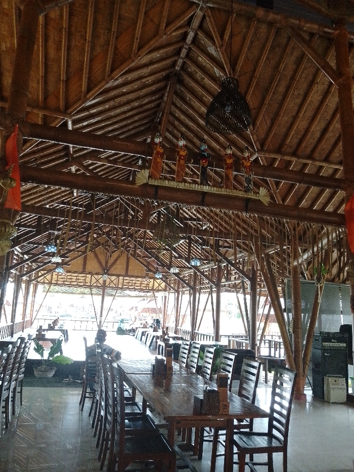 The image depicts the interior of a large, open-sided restaurant or dining area located in Pabelan, Indonesia. The structure features a high, sloped ceiling constructed from interwoven bamboo and wooden beams, supported by numerous vertical and diagonal dark wooden posts. The floor is covered with light-colored tiles. Multiple rectangular wooden tables, each seating several individuals, are arranged in rows with matching wooden chairs. Each visible table has a wooden napkin dispenser and a small potted plant. A decorative display featuring five traditional Indonesian puppet figures, dressed in colorful attire, is mounted on a high horizontal beam, positioned below a large, dark woven pendant lamp. Further decorative elements, including other suspended items and a red fabric banner on the left, are present. One individual, wearing a striped shirt, is seated at a table in the mid-ground, facing away from the camera. Several other indistinct figures are visible seated at tables in the brighter background area. On the far right, a stack of audio equipment, including a large dark speaker cabinet, rests on the floor.