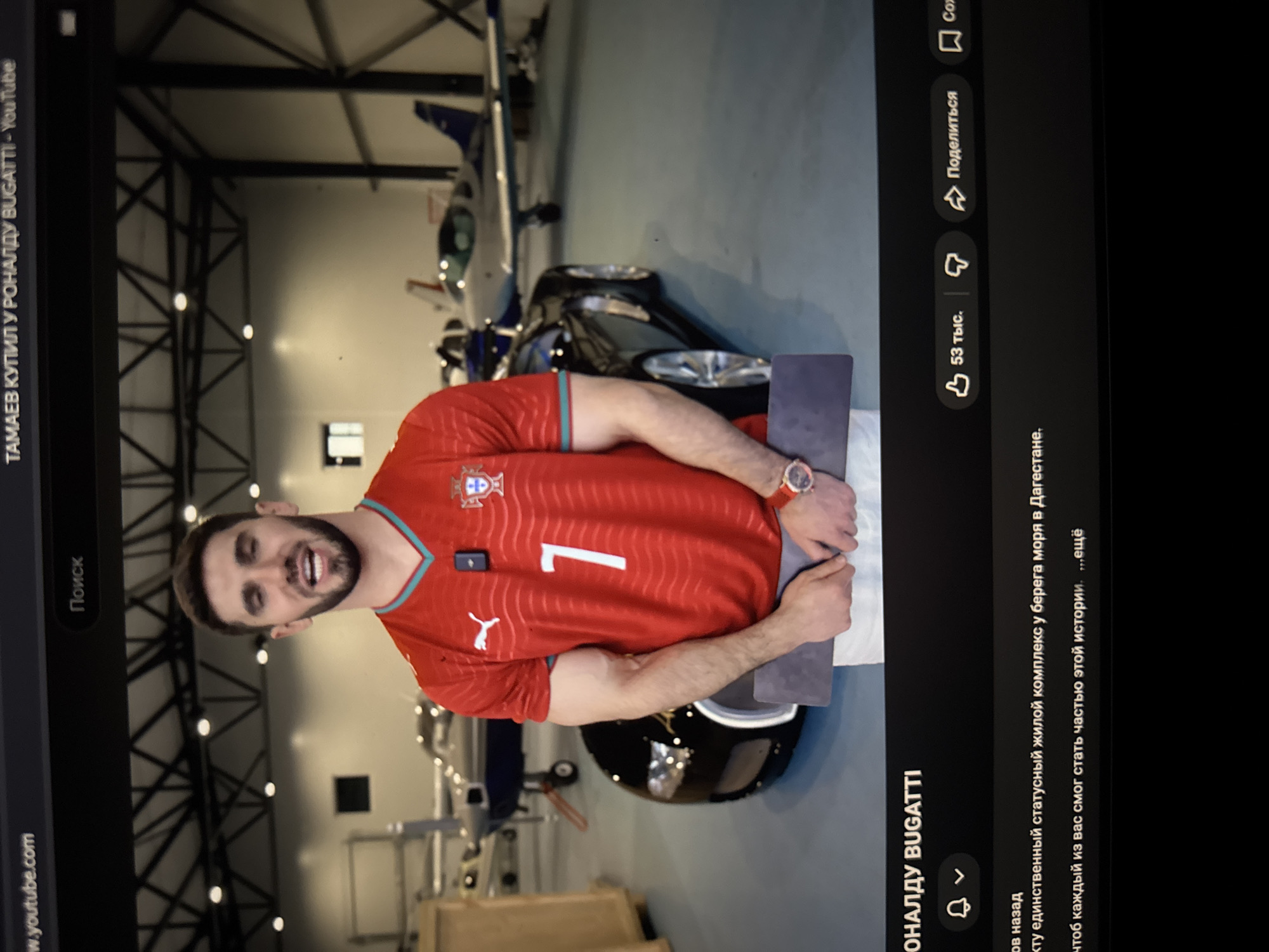 The image shows a screenshot of a YouTube video. In the foreground, a man is standing and looking at the camera. He is wearing a red soccer jersey with the number 7 on it, white shorts, and a red watch. He is holding a dark gray rectangular object in his hands. Behind him, there is a black sports car and several small airplanes in what appears to be a hangar or an aircraft hangar. The background is a bit blurred, but the overall setting seems to be an indoor space with industrial elements like metal beams and lights on the ceiling. The YouTube interface elements are visible at the top and bottom of the screenshot, including the video title "ТАМАЕВ КУПИЛ У РОНАЛДУ BUGATTI - YouTube" and playback controls. The text below the video shows Russian language labels like "Поиск" (Search), "53 тыс." (53 thousand), "Поделиться" (Share), and other video description text. There is no visual information in the image to suggest the location is the city of Демихово, Russia.
