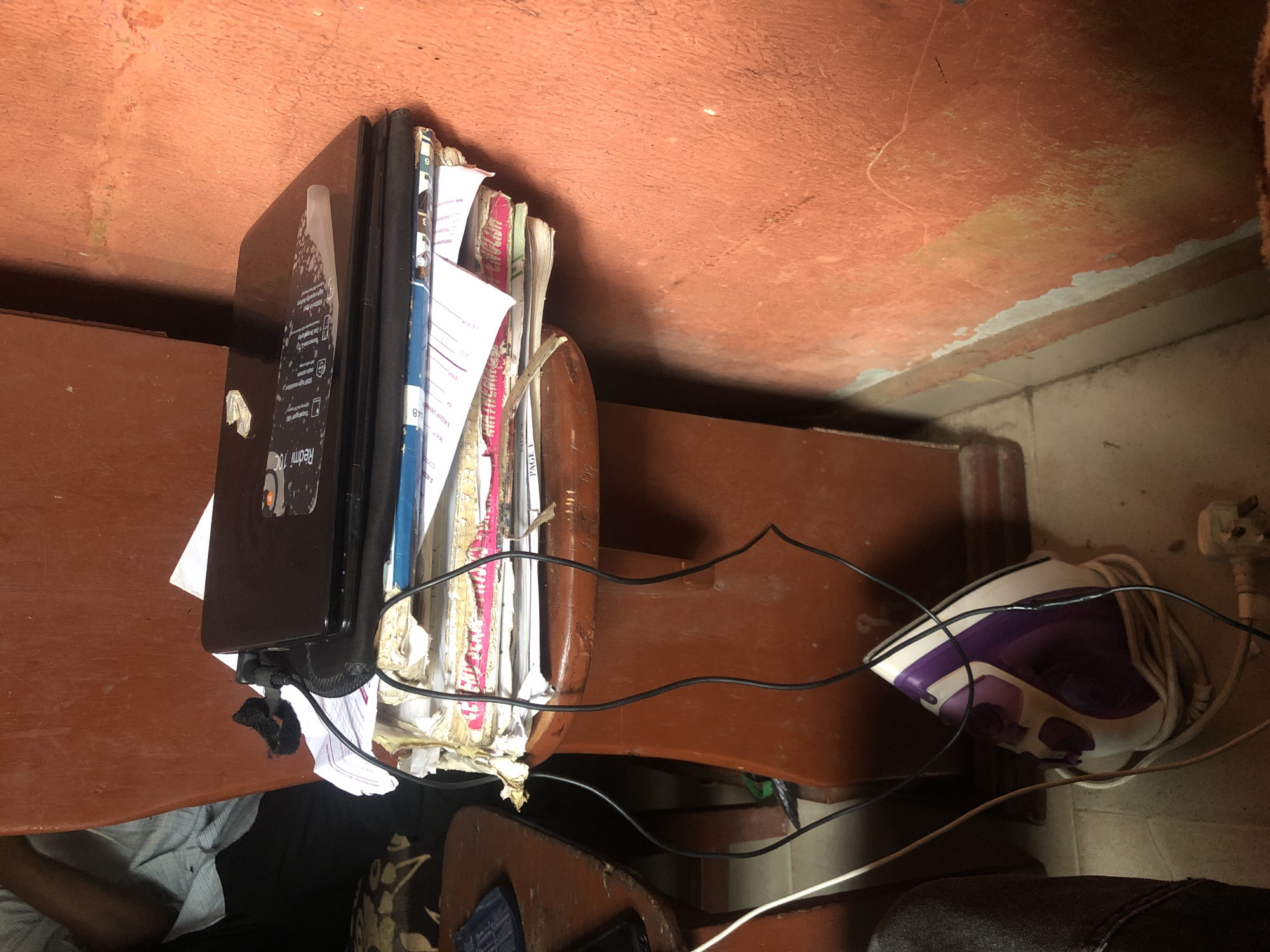 The image depicts a cluttered desk in an indoor setting. A closed black laptop sits on top of a stack of papers and books. The desk is made of brown wood. An iron is located on the floor, with its electrical cord tangled around the desk legs. A person wearing a light blue shirt is partially visible in the lower left corner of the image. The walls are painted in a reddish-brown color. The location is Unknown, Unknown.