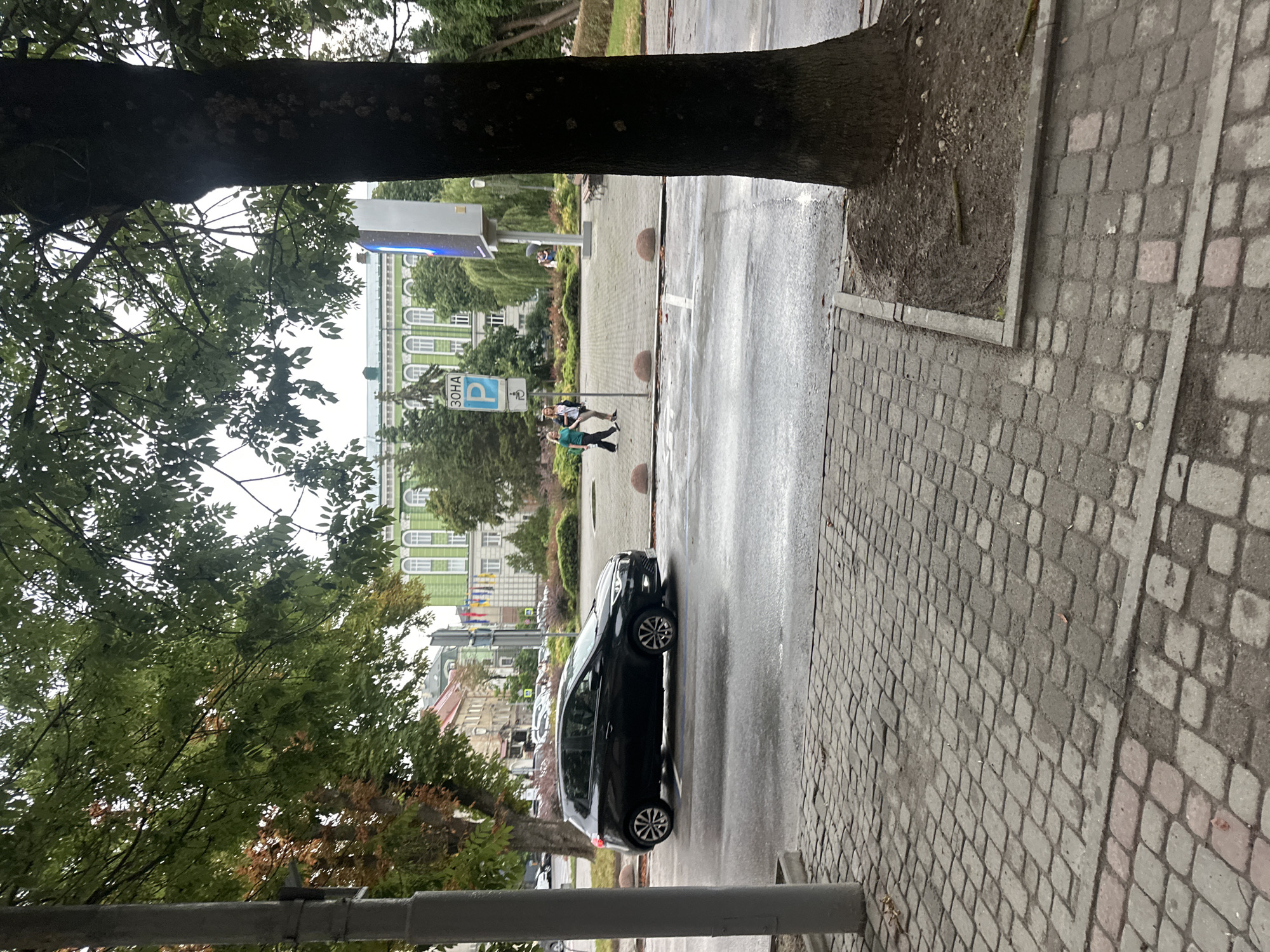 The image depicts a street scene in Ternopil, Ukraine, likely near the Ternopil National Economic University given the presence of a prominent green and white building in the background that resembles its architecture. In the foreground, a paved sidewalk with cobblestone patterns is visible, with a lamppost on the left. A black car is driving on the wet asphalt road. Two individuals are walking on a paved area across the street, near a sign with the letter "P" indicating parking. The environment appears to be a city street with trees, buildings, and other urban elements. The overall atmosphere suggests a cloudy or overcast day due to the diffused lighting and the wetness of the road.