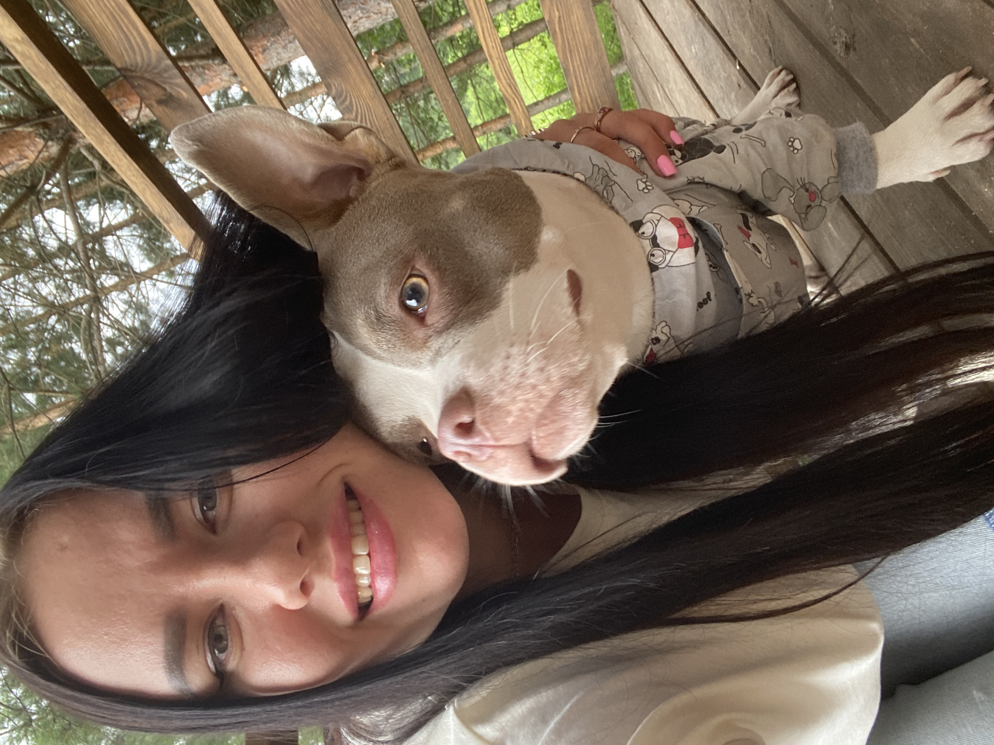 A woman with long dark hair and a light-colored shirt is taking a selfie with a medium-sized dog. The dog is wearing a grey onesie with cartoon characters on it. The dog's head is resting on the woman's shoulder, and they are both looking towards the camera with smiles. They are seated on a wooden porch or deck, with a wooden railing and trees visible in the background. The lighting suggests it is daytime.