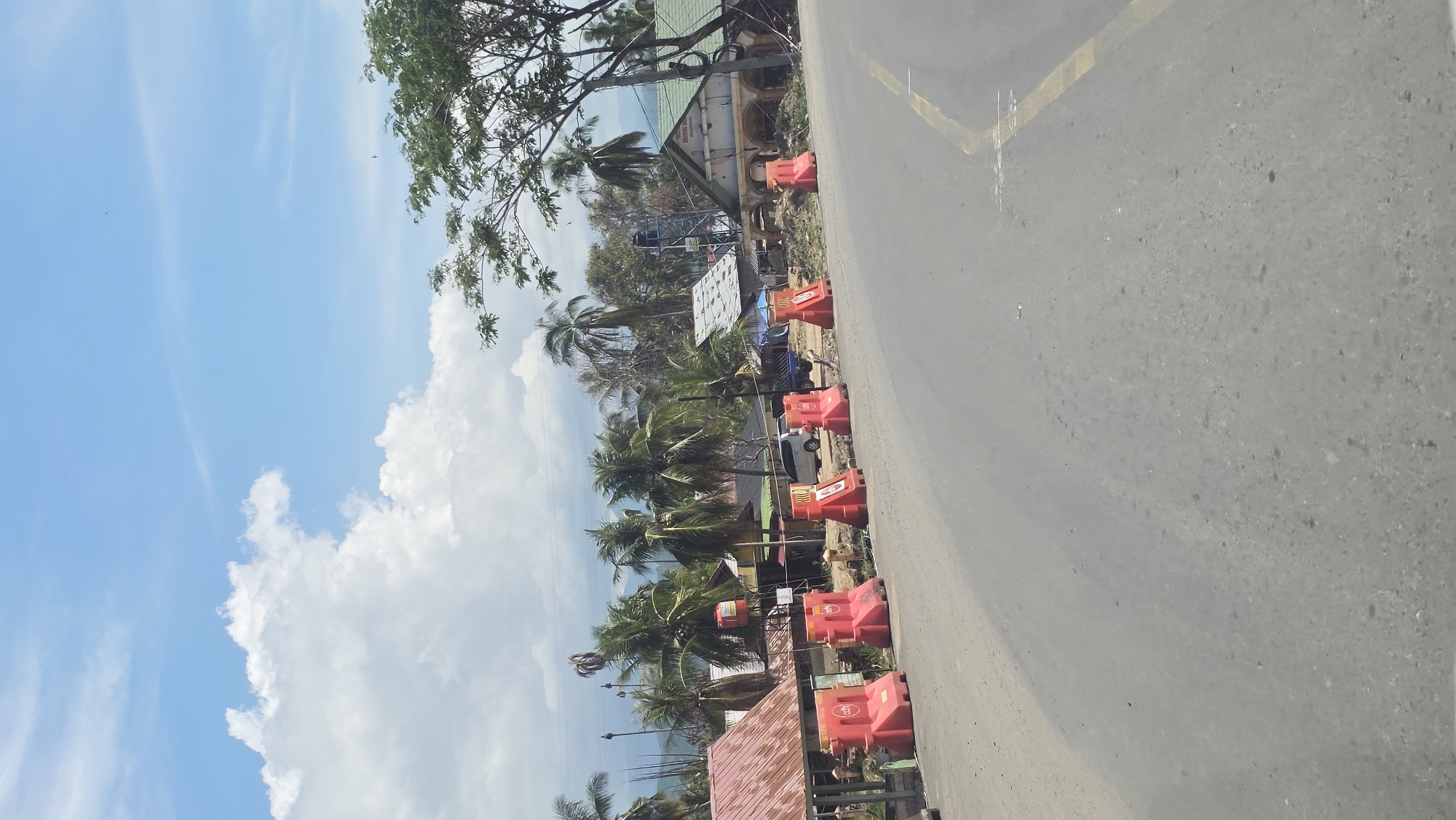 The image shows a road in Manyang Cut, Indonesia, with red and orange safety barriers lining the roadside. Palm trees and various other trees are visible, indicating a tropical environment. The sky is blue with scattered white clouds. A body of water can be glimpsed through the trees. There are buildings of various sizes, some with rooftop solar panels. A few parked vehicles can be seen amongst the trees.