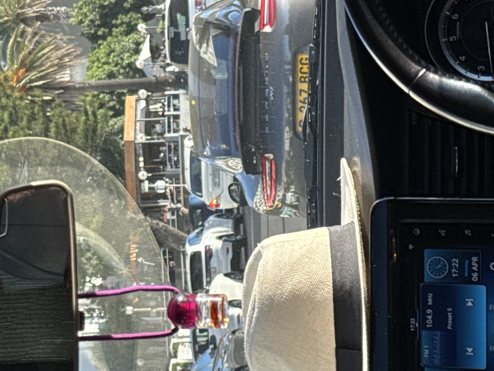 The image appears to have been taken from inside a car, possibly in traffic, in Cape Town, South Africa. The view through the windshield reveals a street lined with parked cars, people, a palm tree, and what seems to be a building or structure. The rear of a dark-colored car with the license plate "G 367 BCG" is visible directly in front of the car. Inside the car, a hat is resting on the dashboard next to the car's infotainment screen, which displays the time as 17:22 (5:22 PM) on Monday, April 6th, and the radio station tuned to 104.9 MHz, preset 5. A cell phone with a pink accessory is attached to the rearview mirror. The dashboard and steering wheel are partially visible, suggesting the make/model of the car. The abundance of cars in the image suggests this might have been taken in a busy part of Cape Town, close to a tourist destination or urban area.