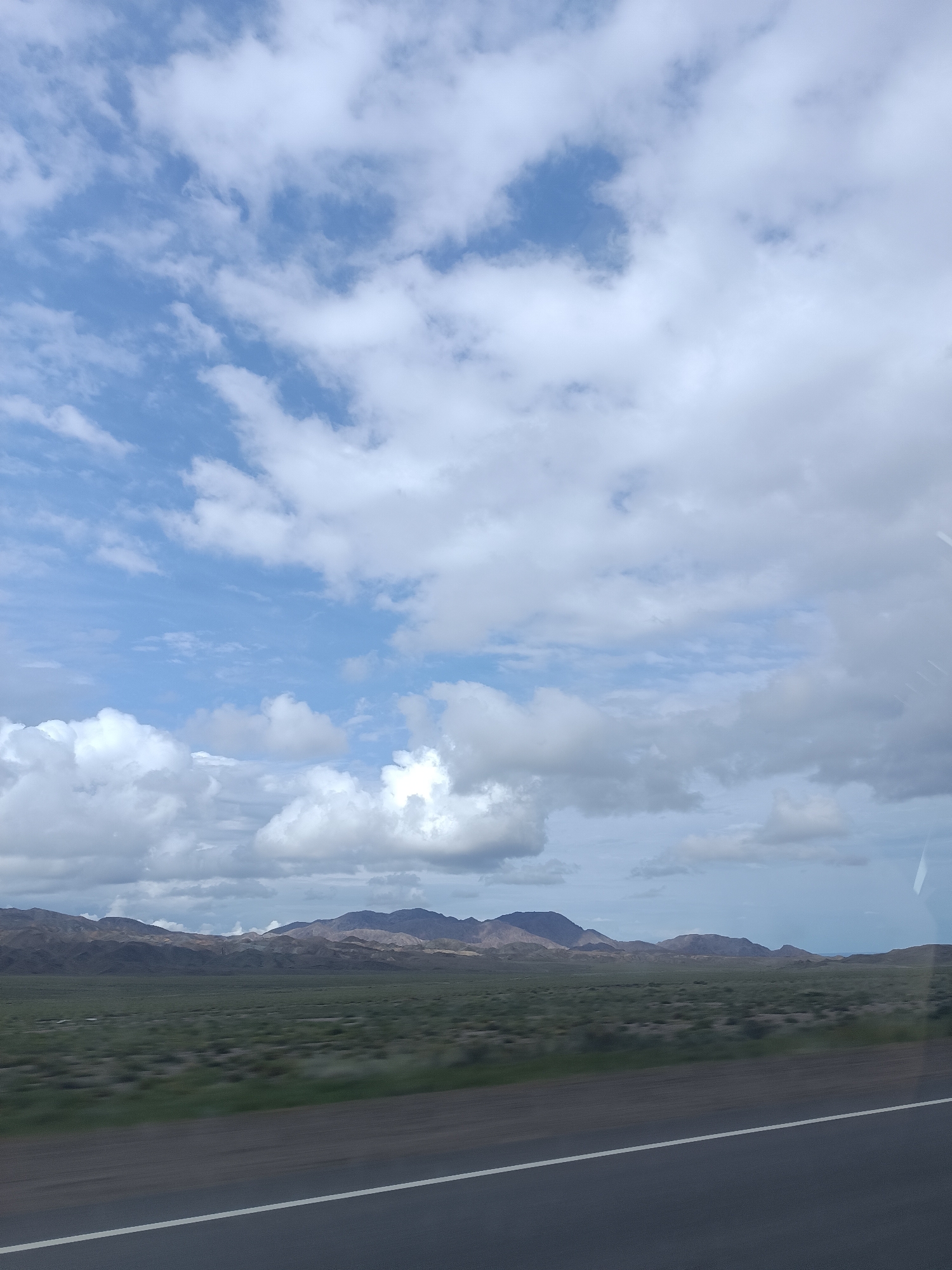 The image shows a landscape with mountains in the background and a vast plain in the foreground. The sky is filled with clouds, some of which are white and fluffy, while others are darker and more ominous. The plain is covered in low-lying vegetation, and there is a road in the foreground with a white line marking its edge. The lighting suggests it is daytime, possibly late afternoon, given the diffused light. The scene is likely from a rural or natural area, possibly along a highway. There are no people or specific objects visible that would pinpoint an exact location within Sasyk-Bulak, Kyrgyzstan, but the overall scenery is consistent with a mountainous, arid, or semi-arid region.